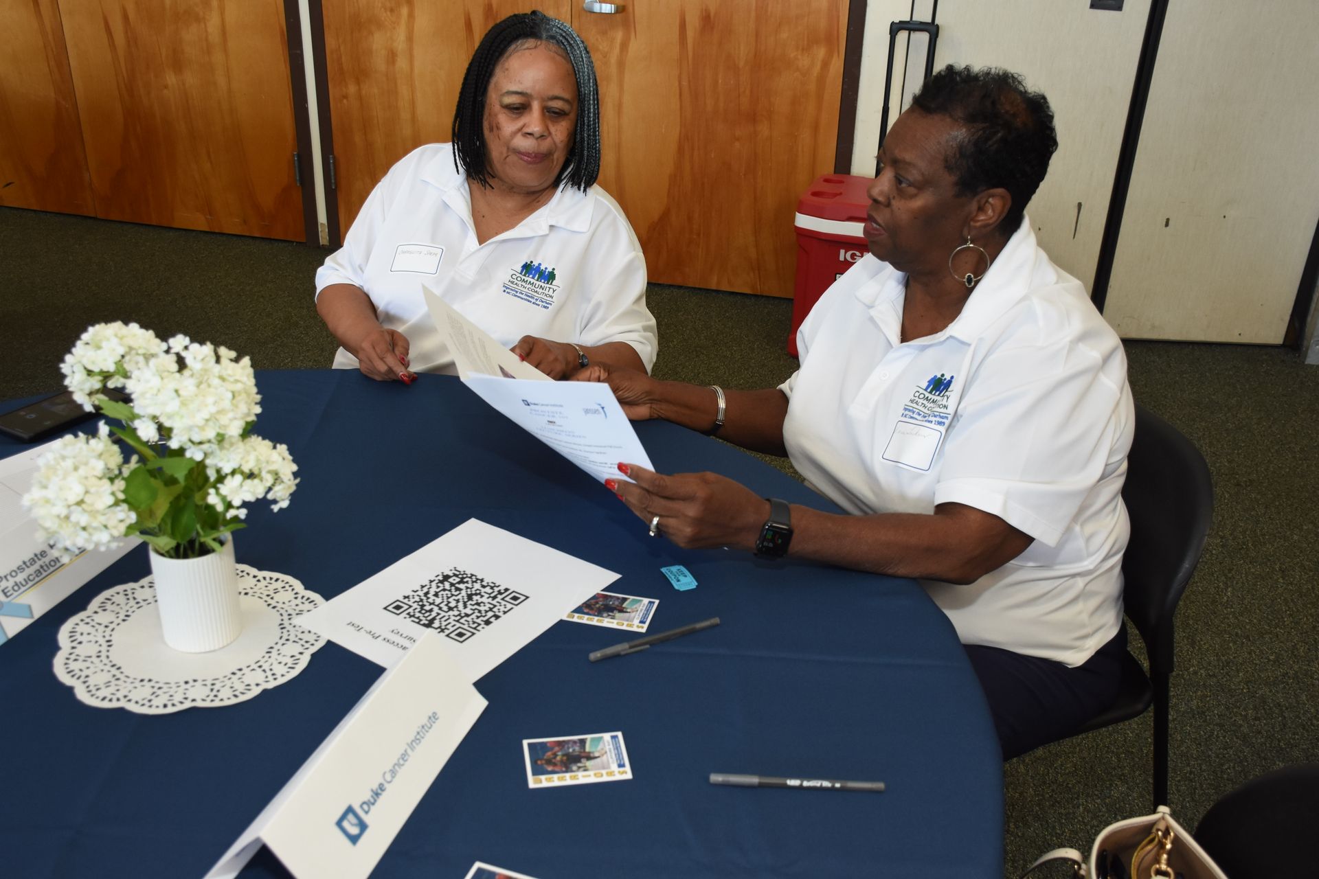 Two women in white shirts with logos sit at a round table, reviewing papers; flowers in a vase sit to the side.