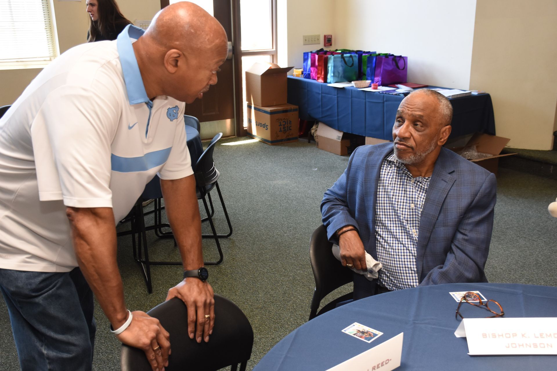 Two men conversing in a room; one standing in a UNC shirt, the other seated in a blue blazer.