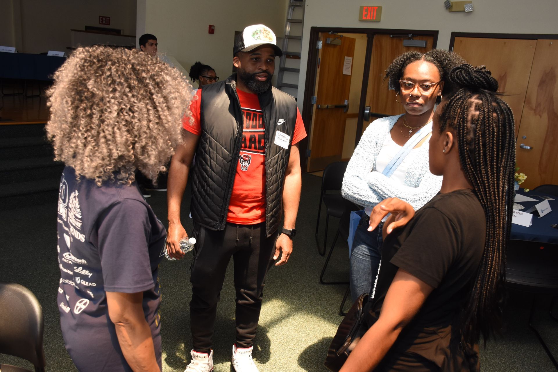 Group of people conversing in a room. Man in vest, hat, and shirt speaks. Others listen and engage.
