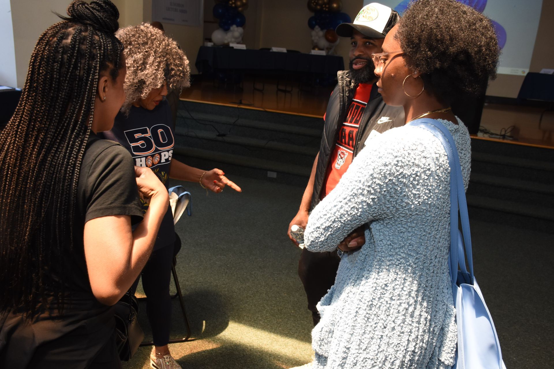 Four people converse in a room with blue decorations. Two women and a man are facing each other, one woman gestures.
