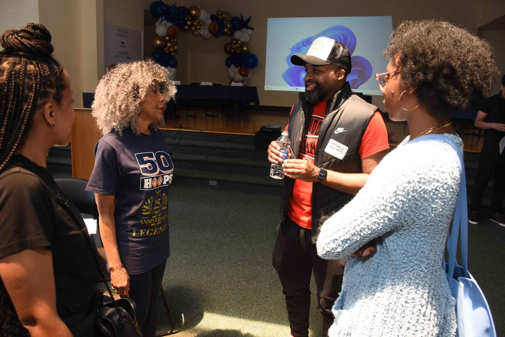Four people conversing indoors, two women facing two others, one man in a hat talking.