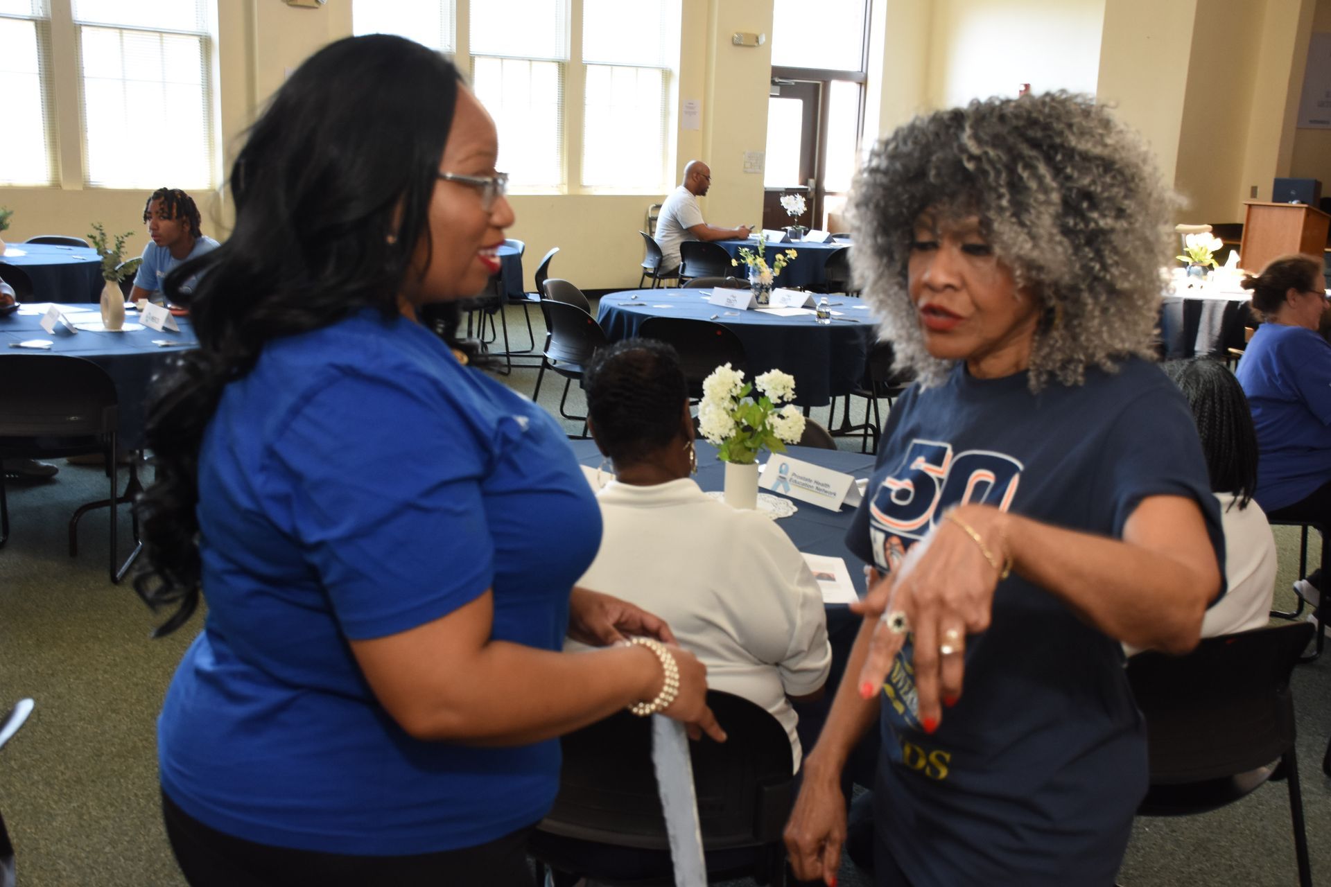 Two women in blue shirts talking at an event; tables and people in the background.