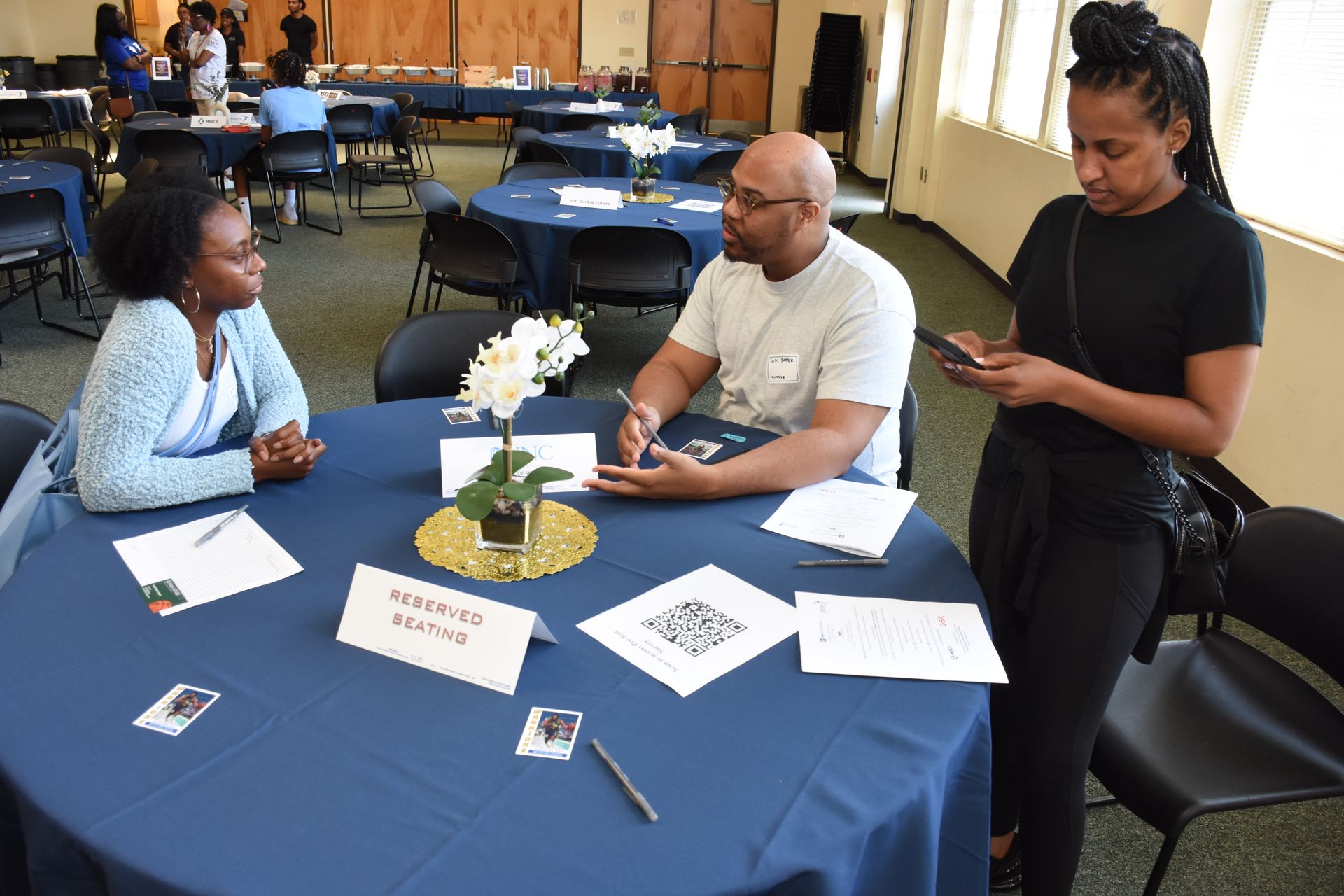 Three people at a table, one looking at phone. Event setting, blue tablecloths, white flowers, name tags.