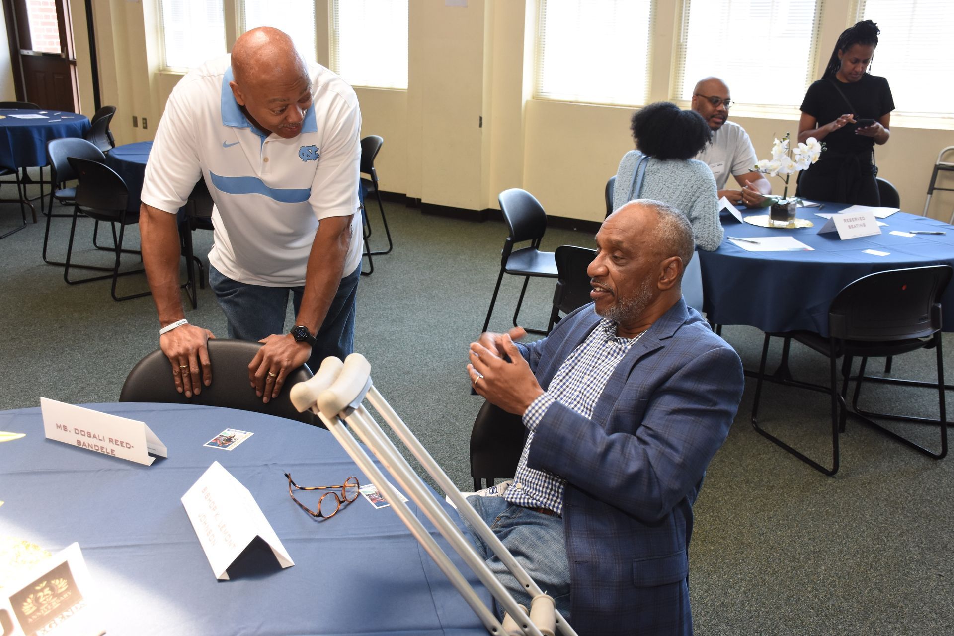 Man in a light blue shirt talking to a man seated with crutches at a table. Other people are seated at tables in a room.
