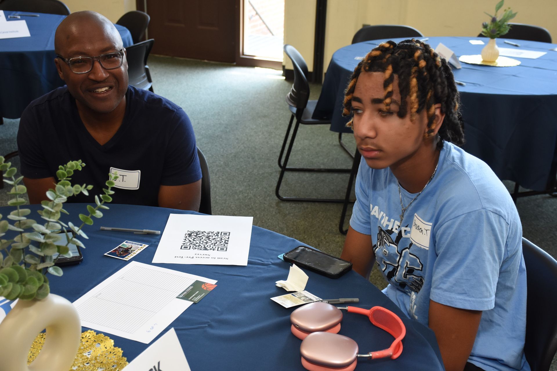 Two people sit at a table in a room with a blue tablecloth. The man smiles and the youth looks forward.