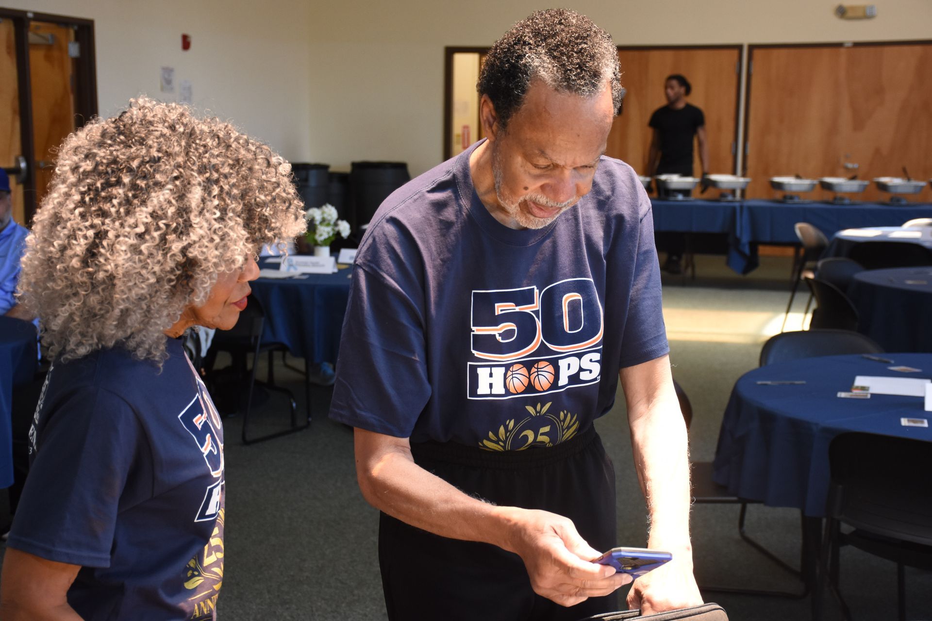 Two people looking at a phone in a room with tables set for a gathering.