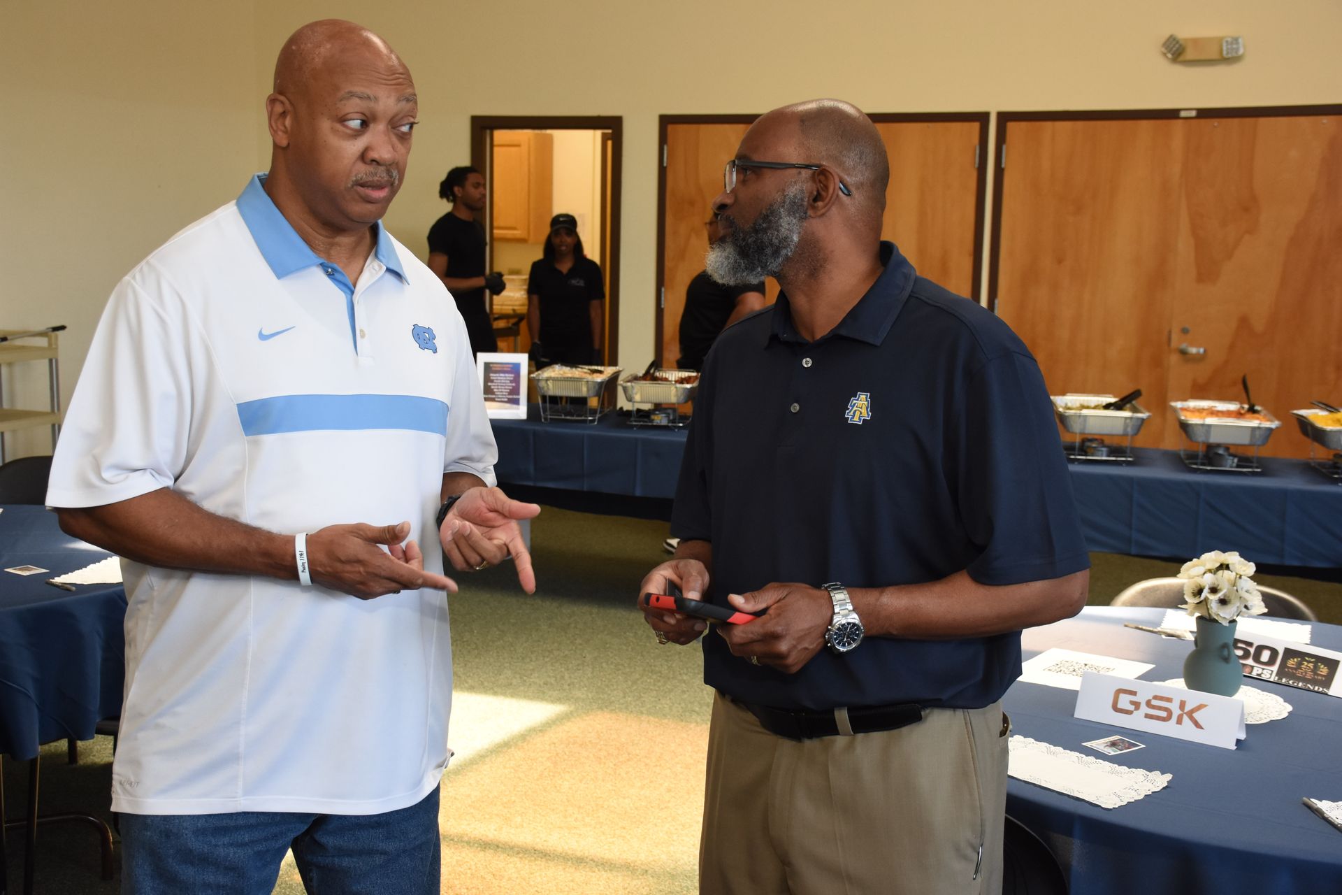 Two men talking at a gathering, near buffet tables. One wears a light blue polo, other in dark blue.