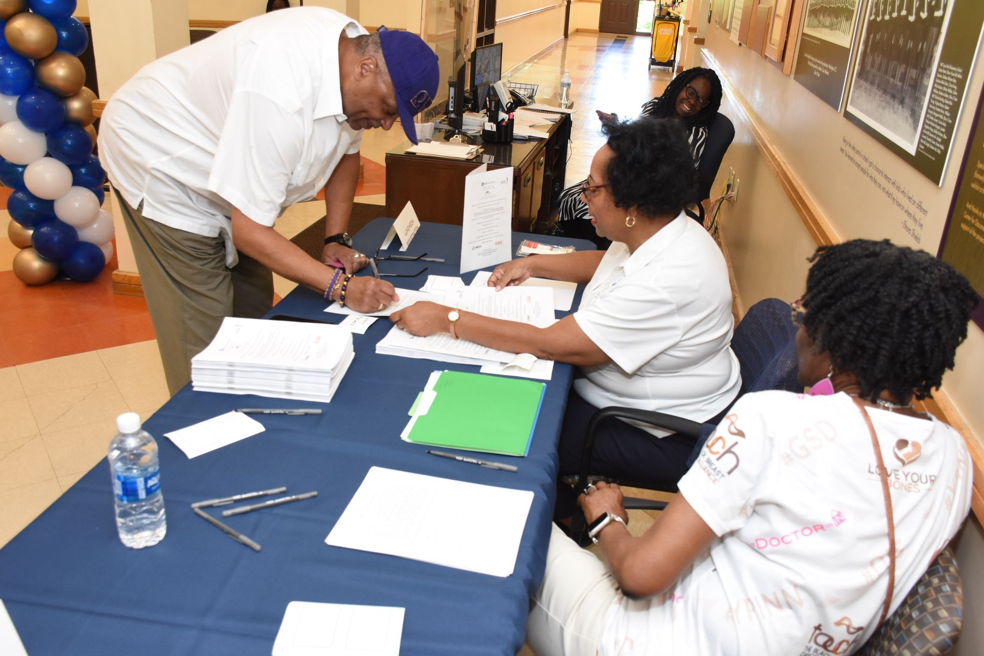 Man signing documents at a table. Two people sit behind the table, a bottle of water and balloons visible in the background.