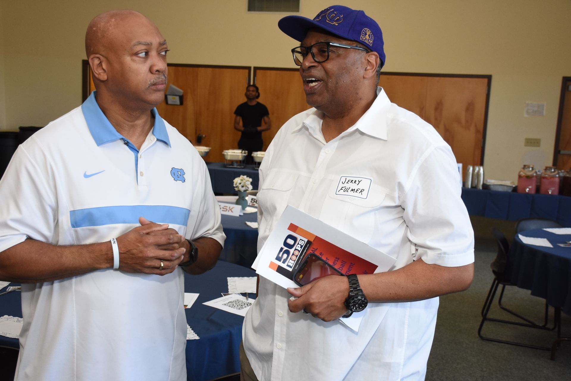 Two men talking indoors; one wears a Carolina blue shirt, the other a white shirt and blue cap, holding a brochure.