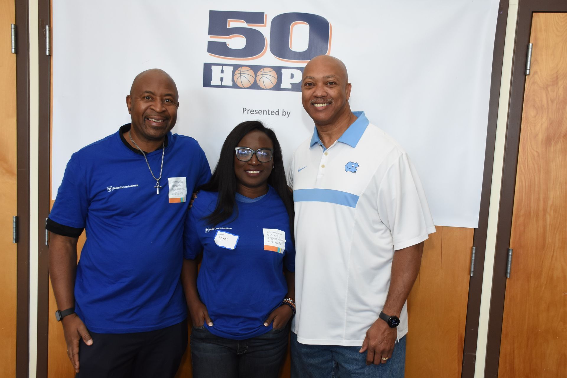 Three people pose in front of a banner. Two men in blue and a woman in the center, smiling.