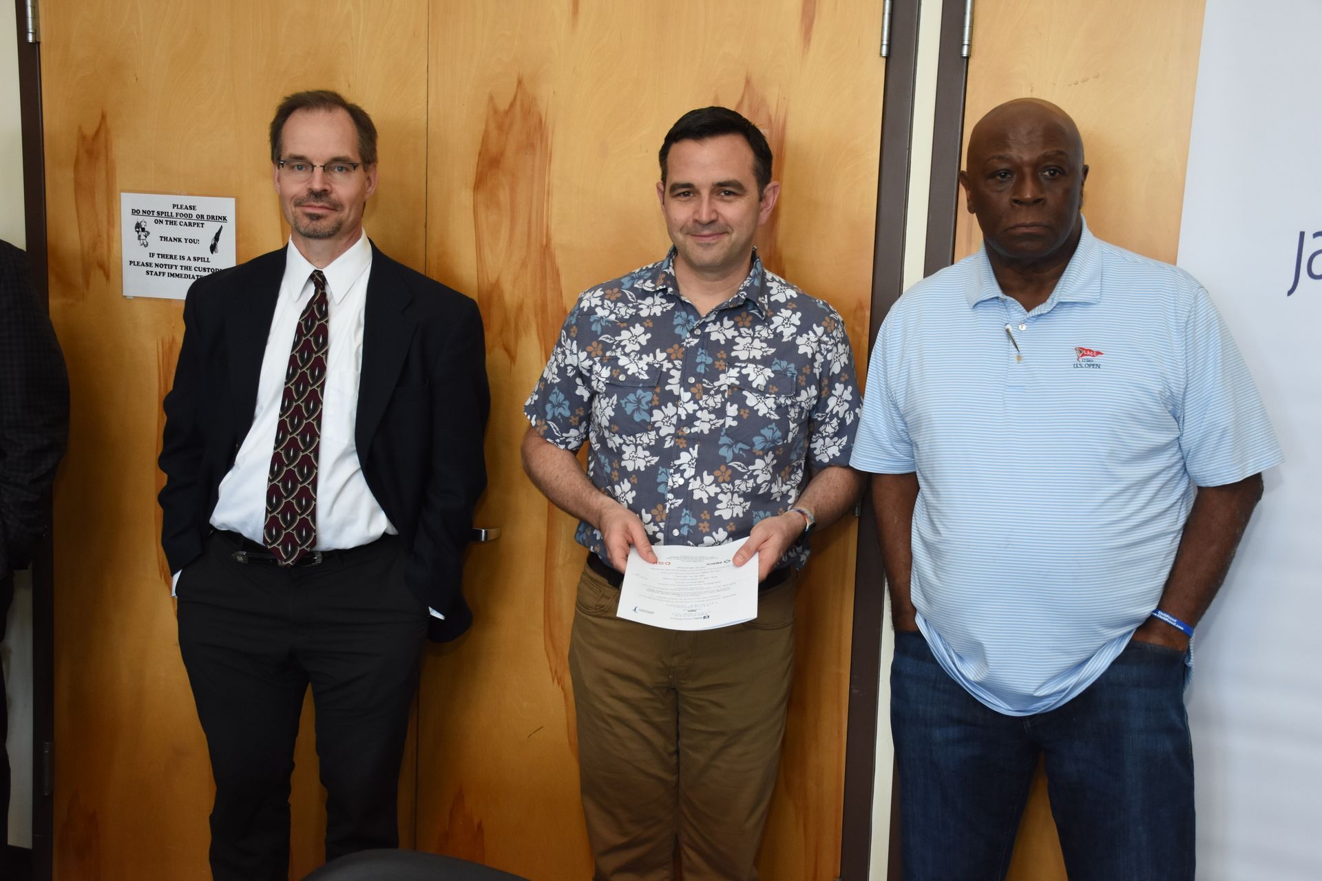 Three men standing in front of a wooden wall. One holds a check, others in suits and a polo.