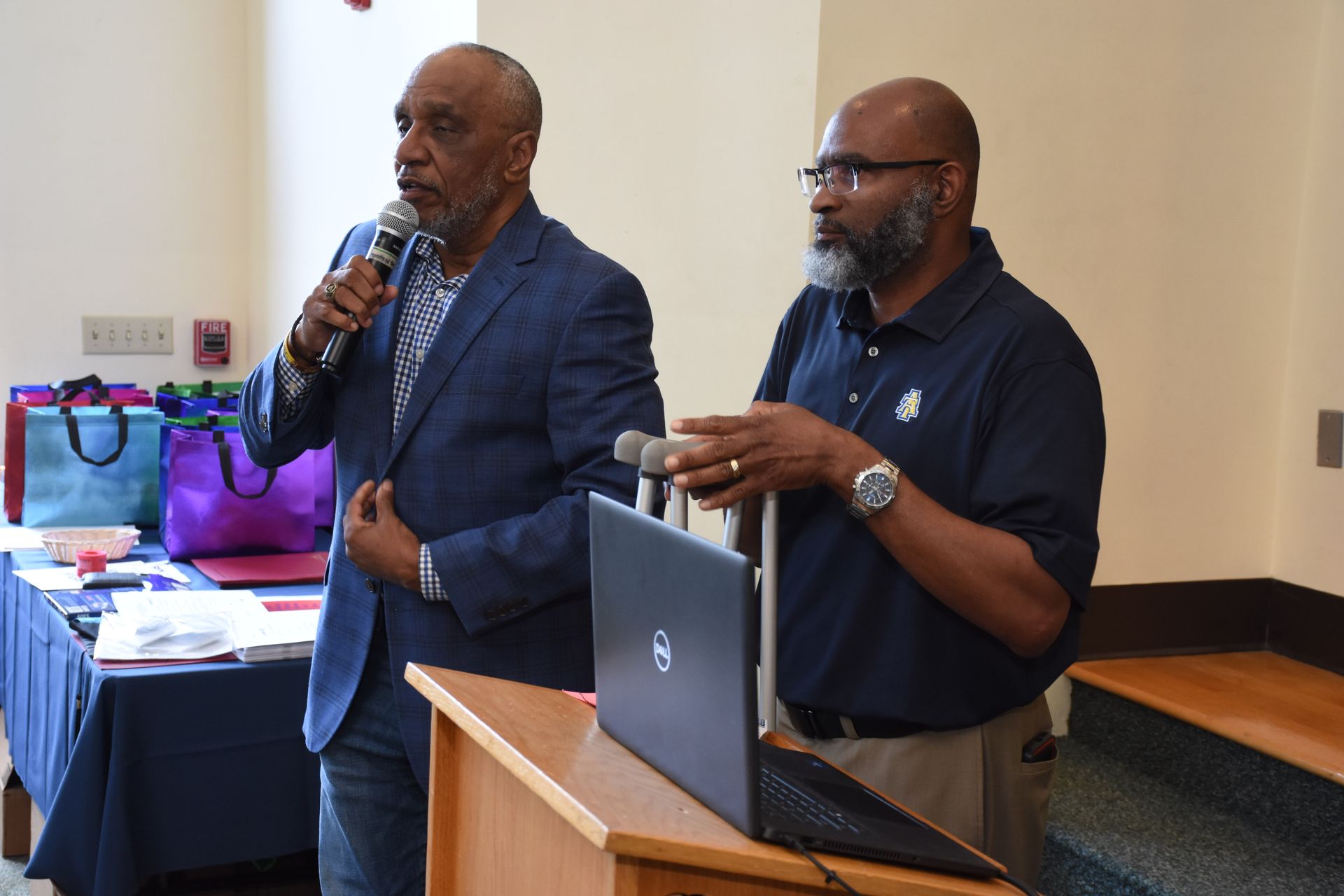 Two men at a lectern: one speaks into a mic, the other holds a mic, laptop in front, table with gifts in the background.