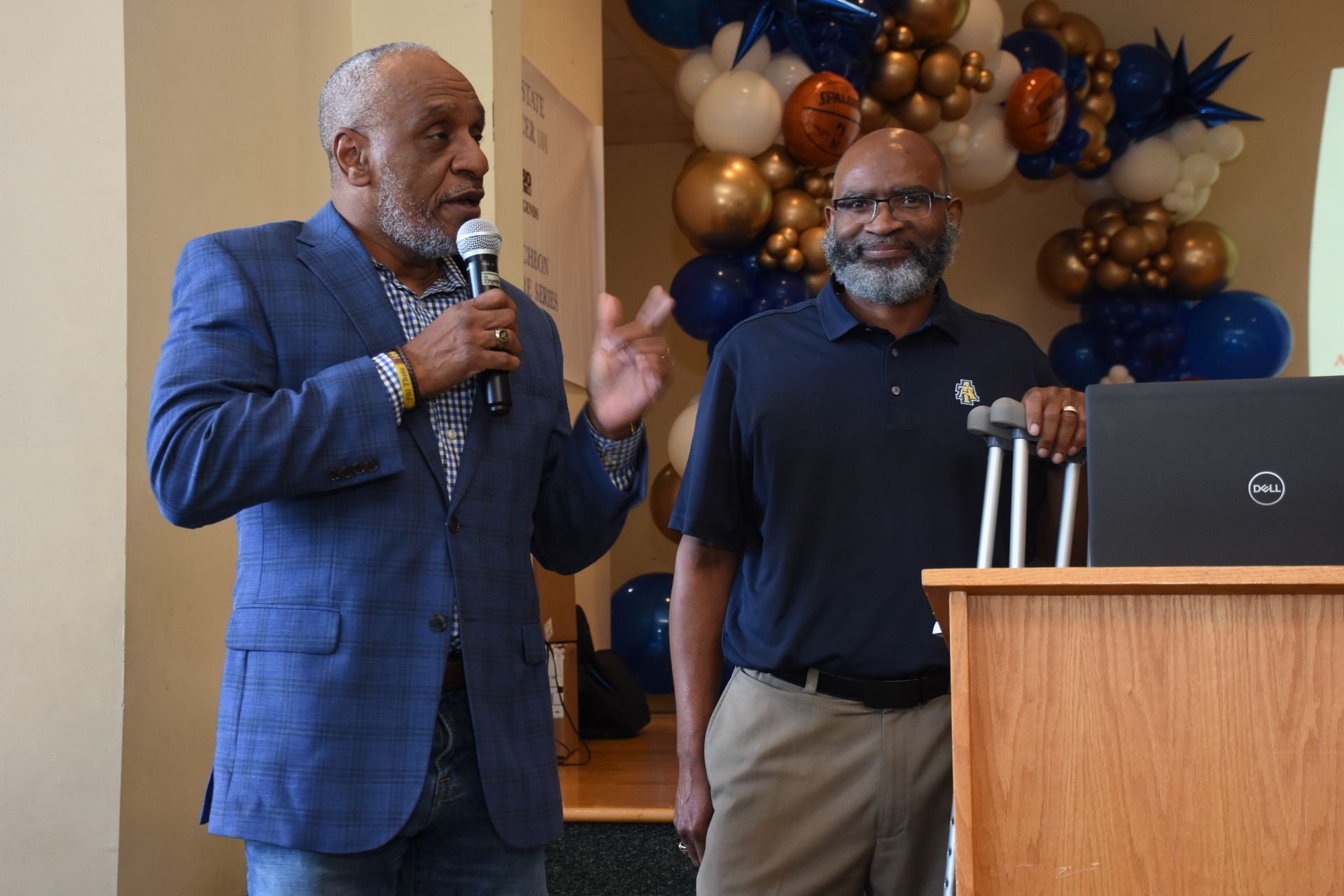 Two men at a podium; one speaks into a mic, the other stands with crutches. Background has balloons.