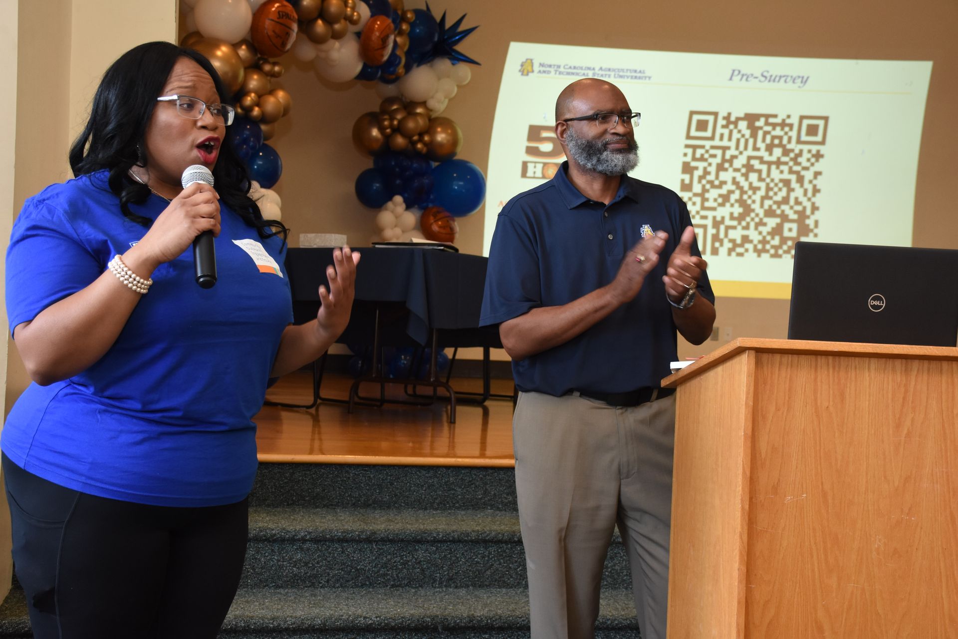 Woman speaking into a microphone, man clapping, at a podium, presentation screen visible.