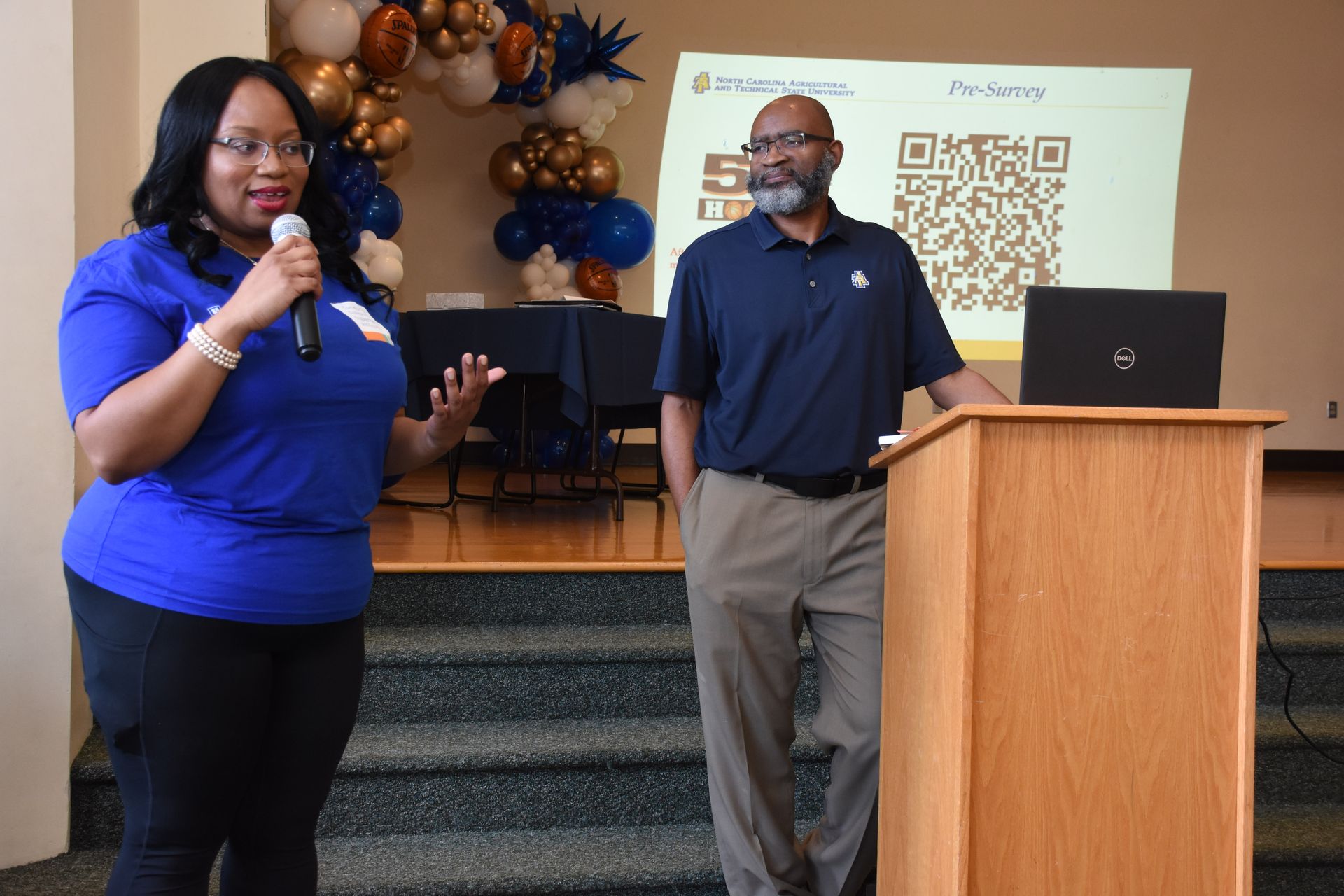 Woman speaking with a microphone, man beside podium; a presentation in a room, balloons, and a QR code visible.