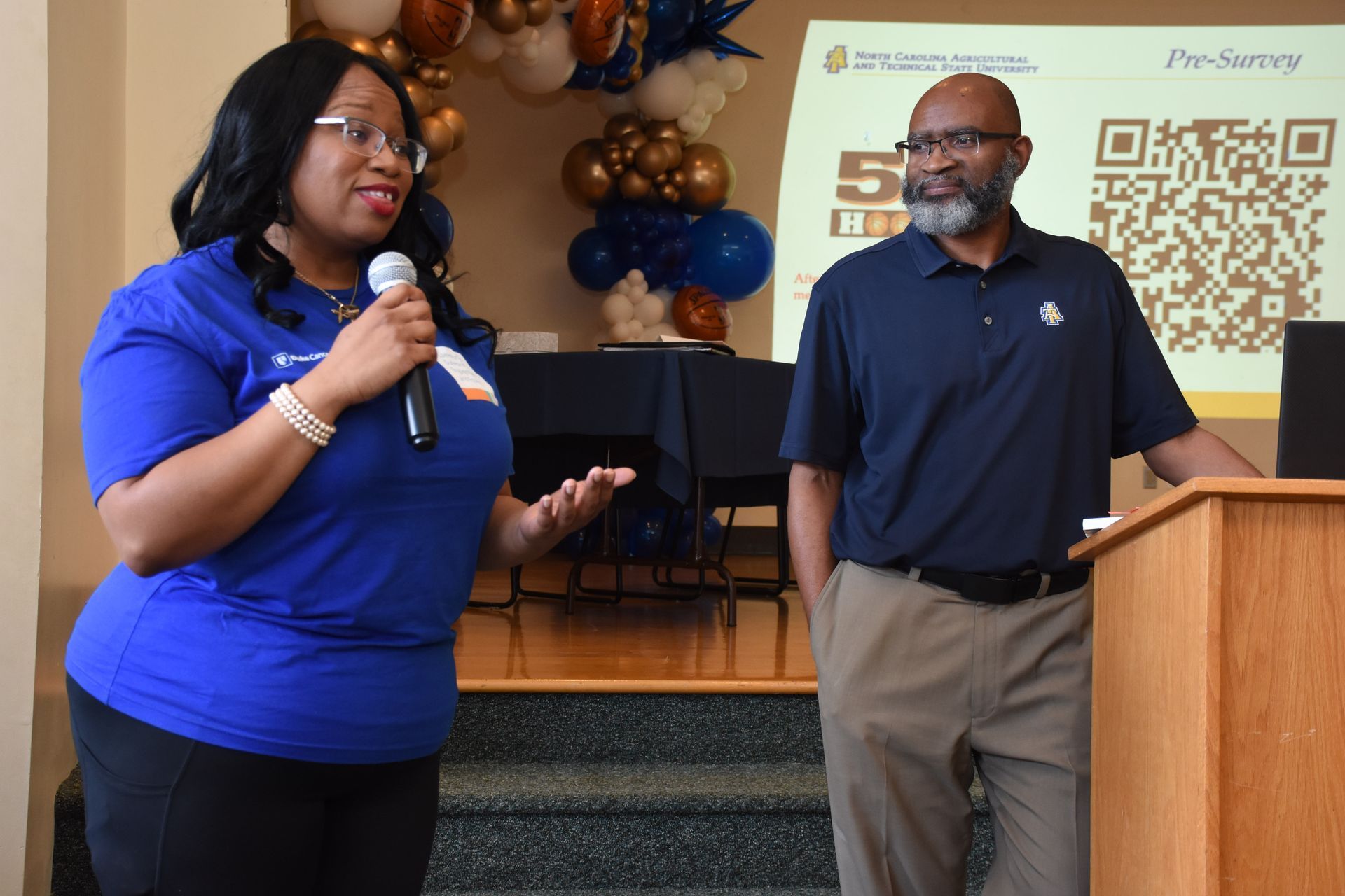 Woman in blue shirt speaks into microphone; man in blue shirt stands by lectern. Balloons decorate background.