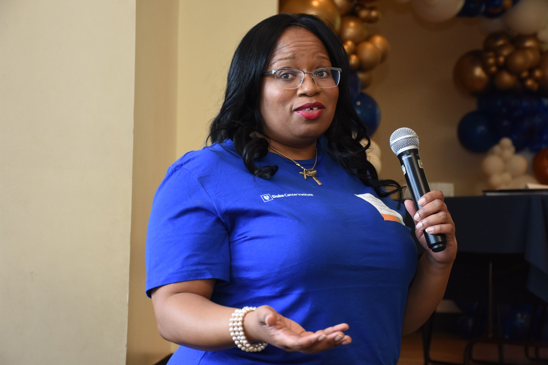 Woman in blue shirt speaking into a microphone, gesturing with her hand at an event.
