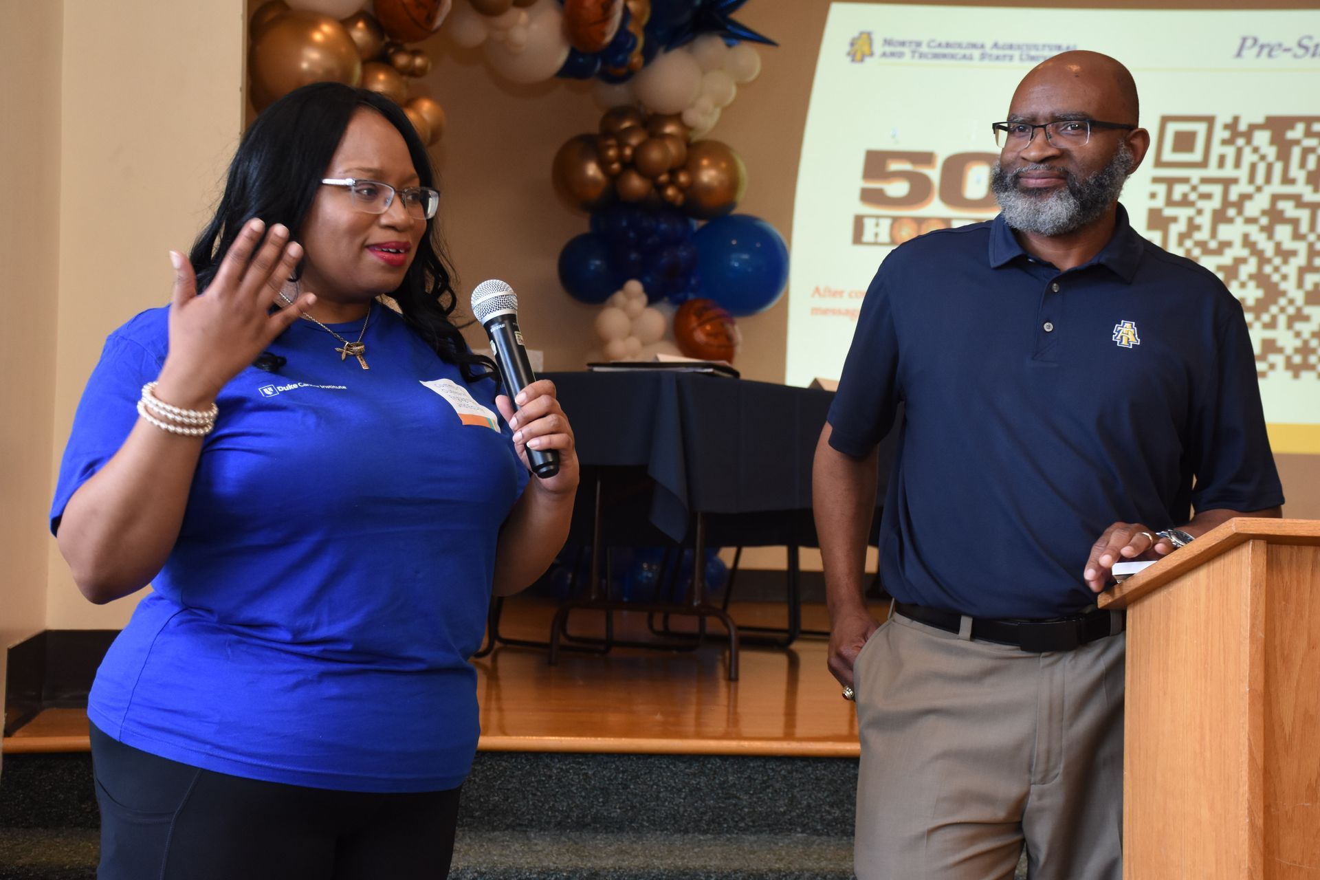 Woman speaking at podium, man beside her. Balloons and screen in the background; blue and gold colors.