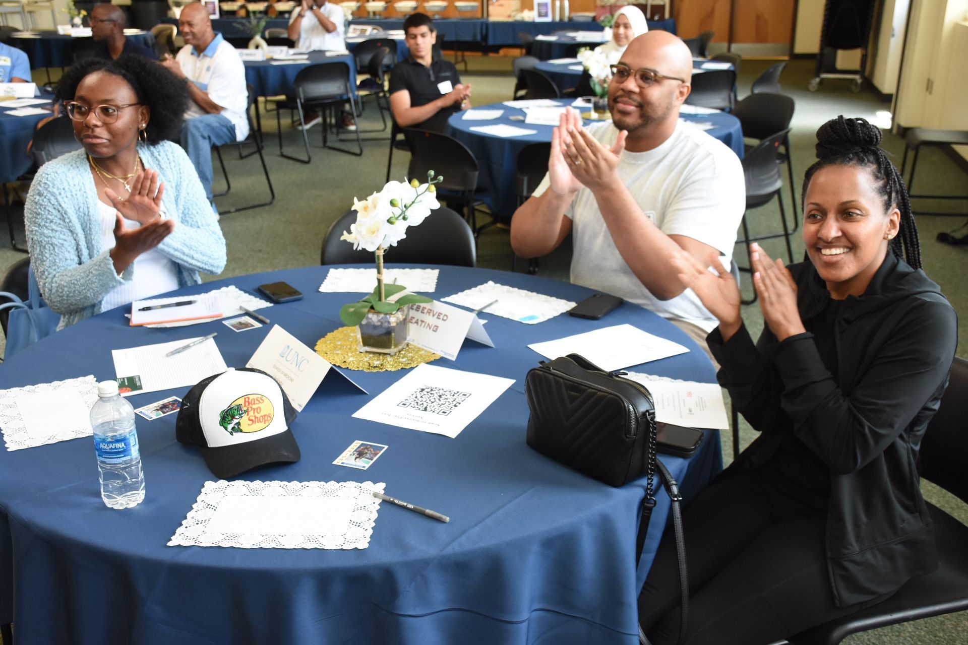 People clapping at a round table during a meeting; blue table with papers, water, hat.