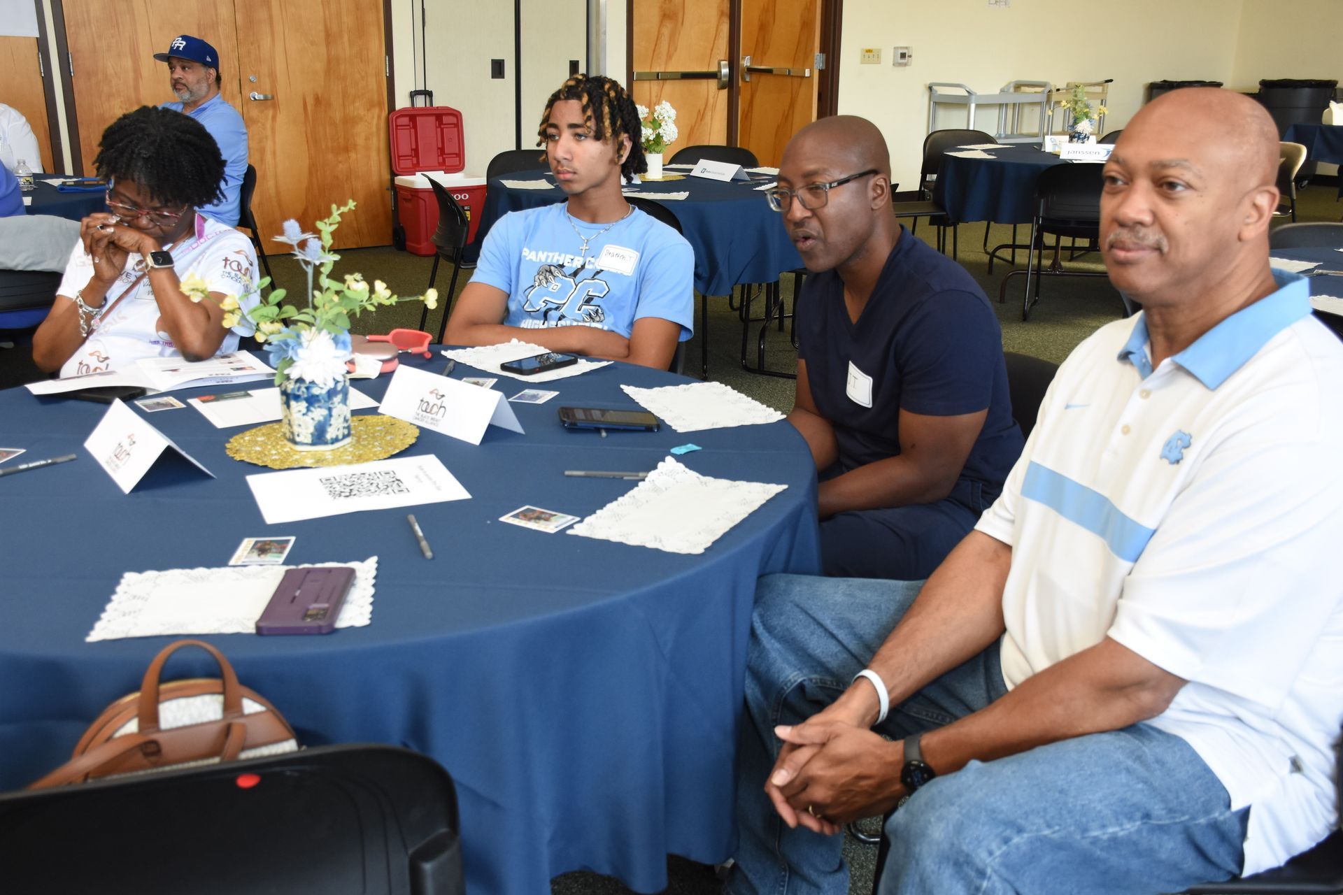 People seated around a blue table in a conference room, listening and observing, some wearing nametags.