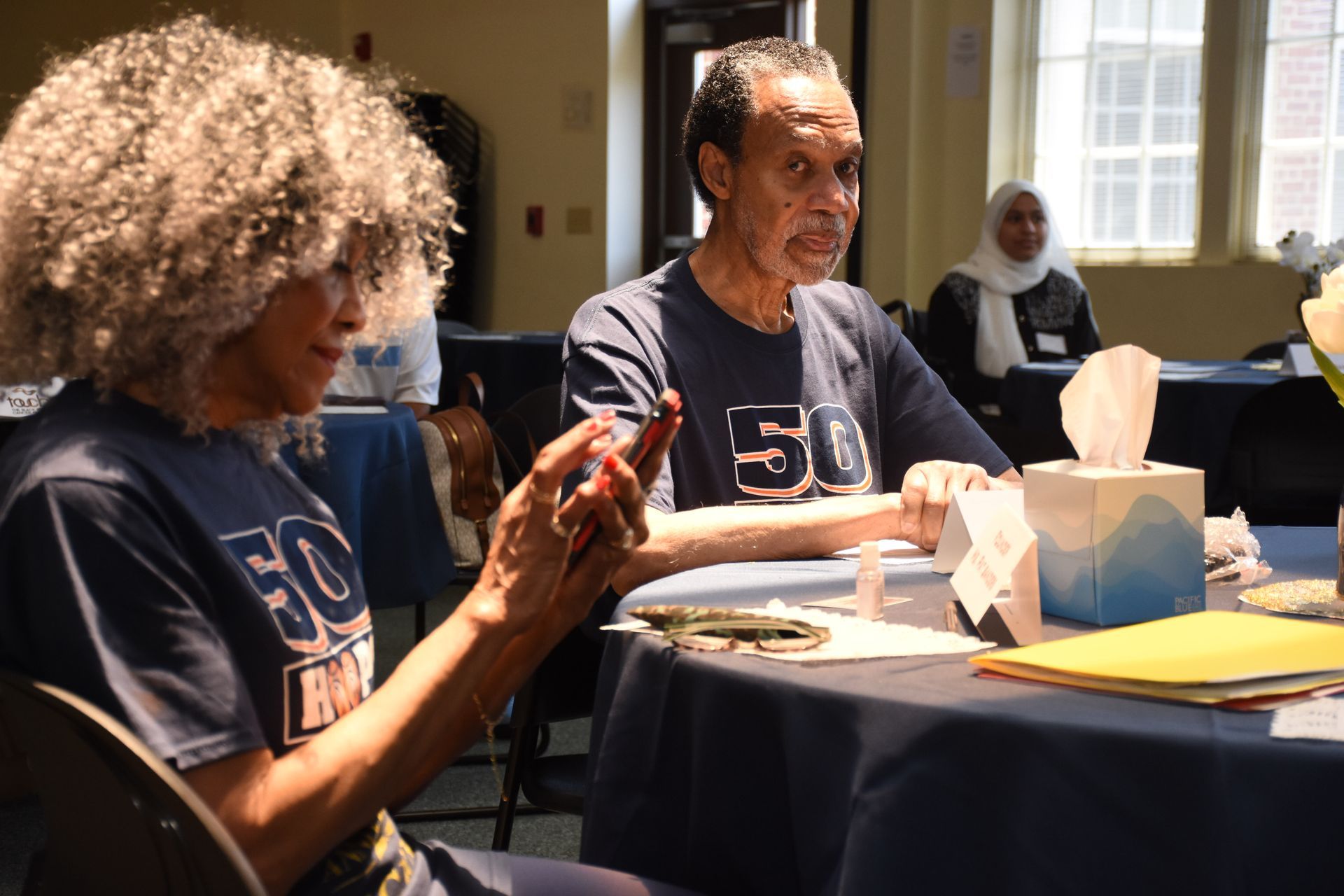 People seated at a table, some wearing matching shirts. One woman is looking at a phone.