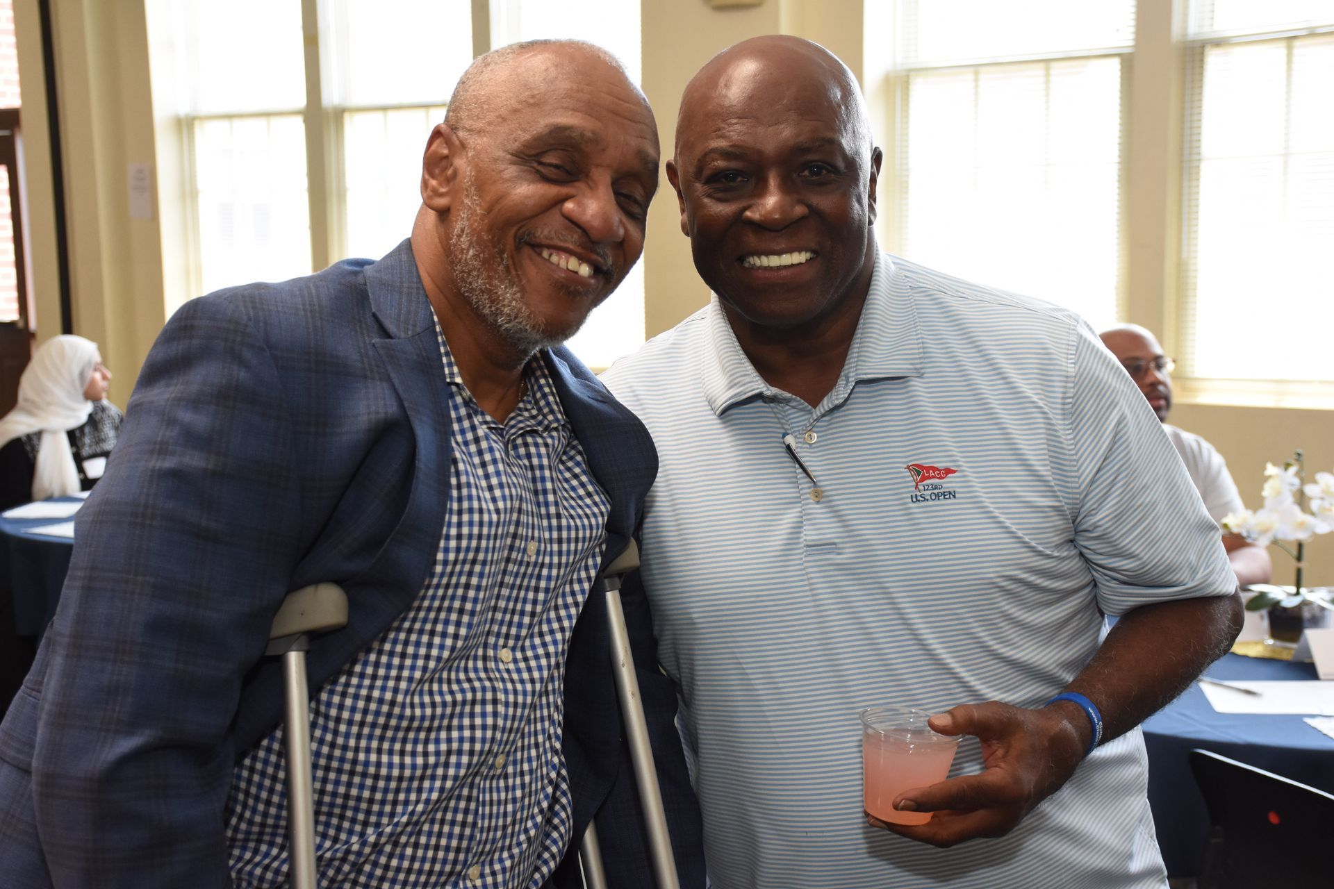 Two smiling men pose for photo. One with crutches, blue blazer. Other in light blue polo holding a drink.