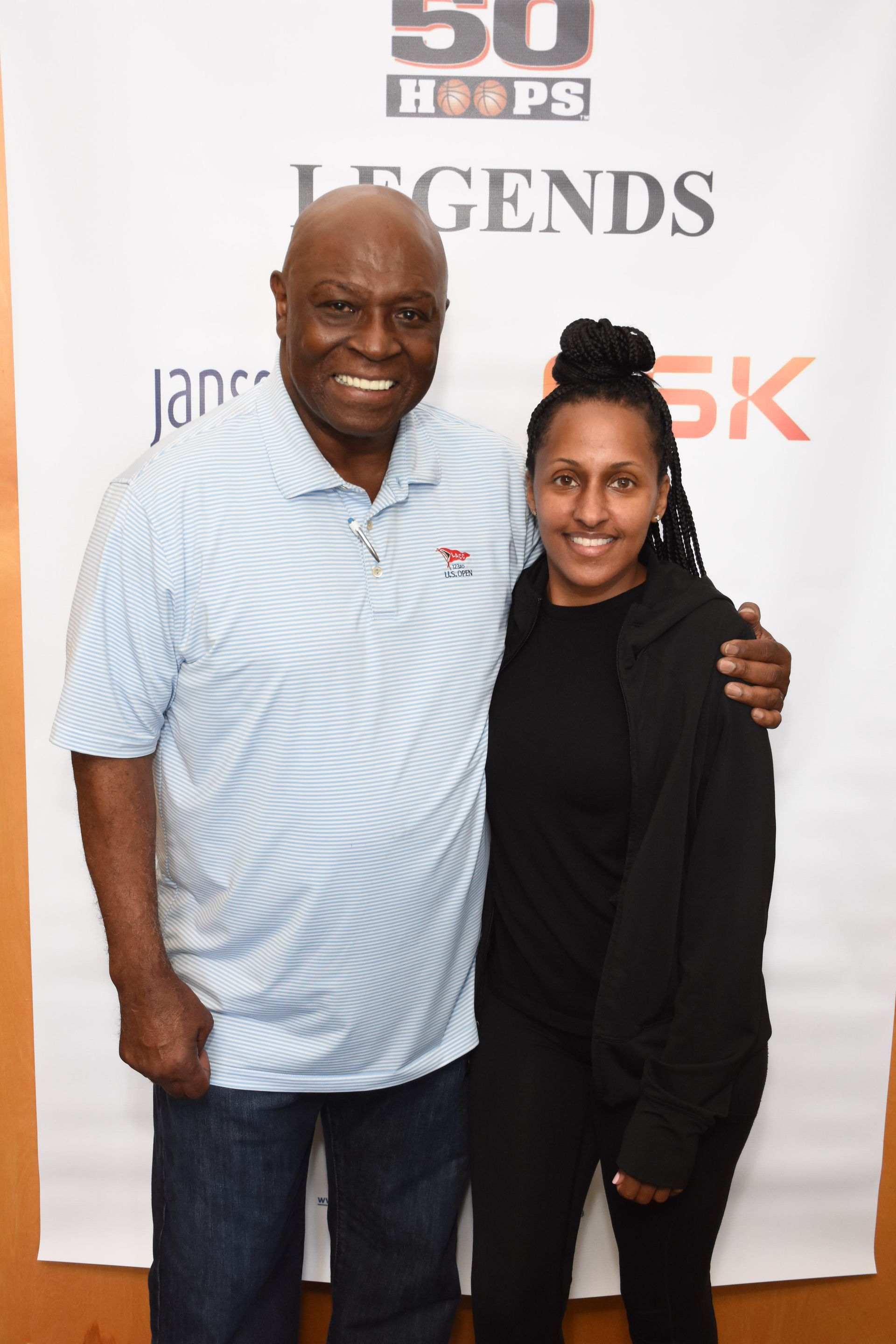 Man in blue shirt and woman in black hoodie pose smiling in front of a sign.