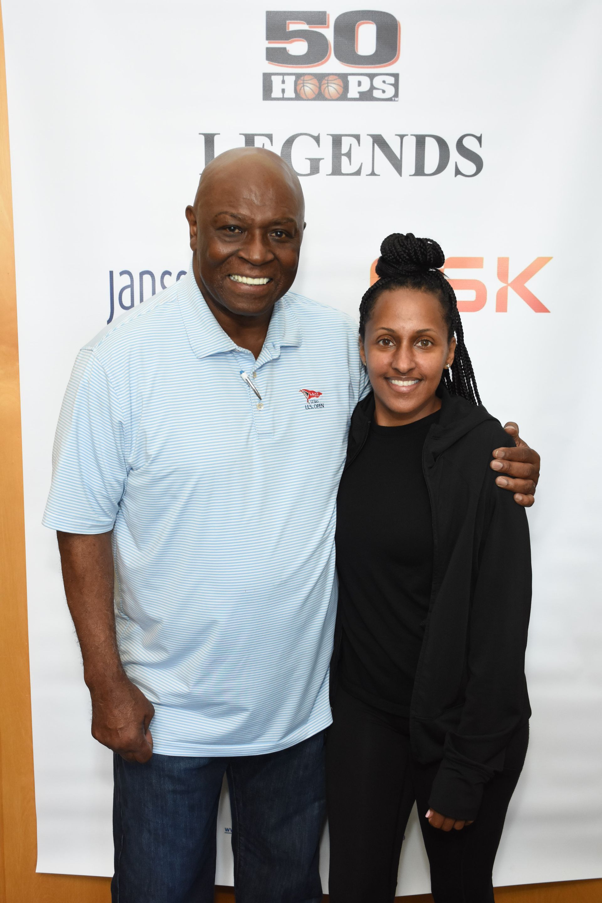 Man in light blue shirt, dark-haired woman posing together at an event. White backdrop with 