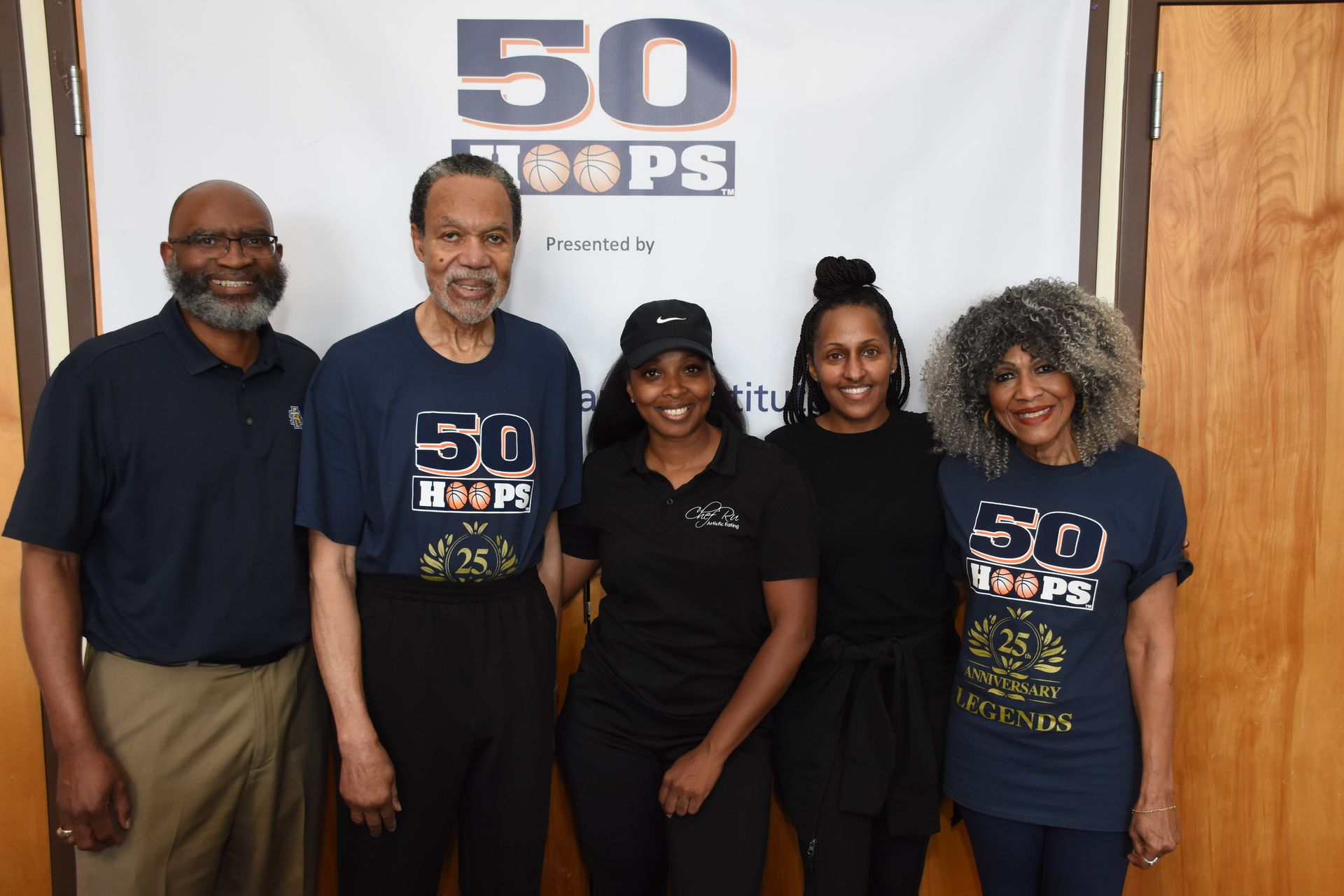 Five people pose in front of a banner. They are wearing “50 Hoops” t-shirts. Setting is indoors.