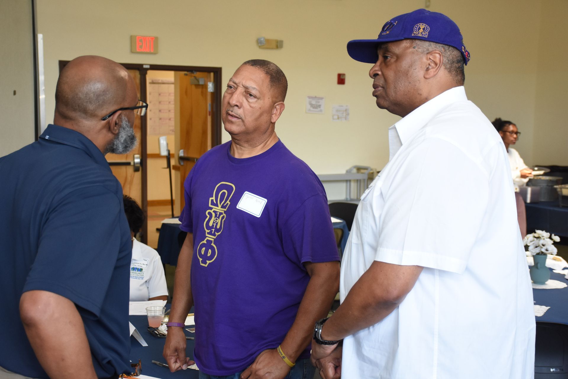 Three men in a room talking. One wears a purple shirt, another a white shirt and cap, and the last a navy shirt.