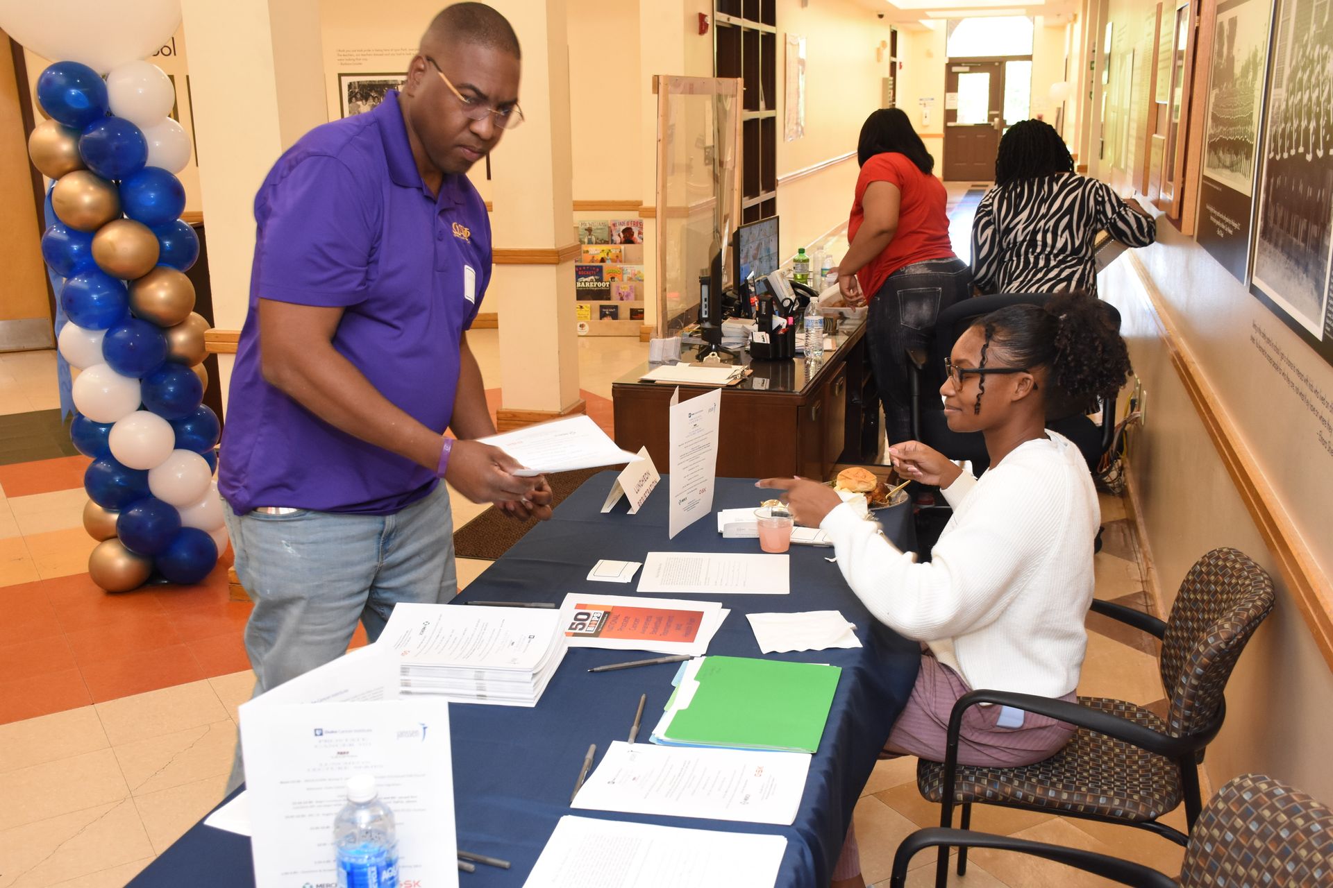 Man in purple shirt reviews papers at a table, woman behind table points. Indoor setting, balloons, people in background.
