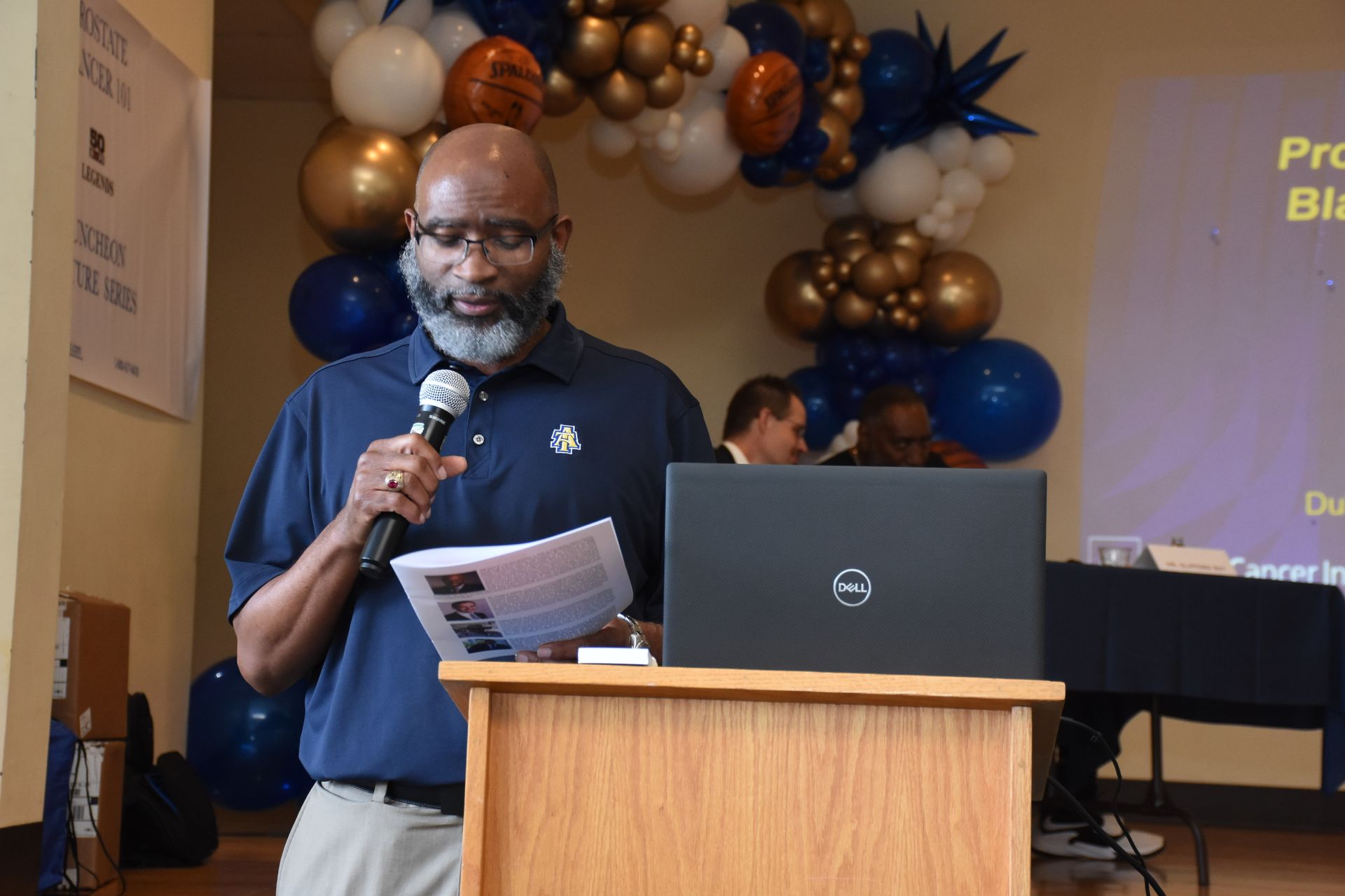 Man speaking at a podium with a microphone and laptop, blue and gold balloons in the background.