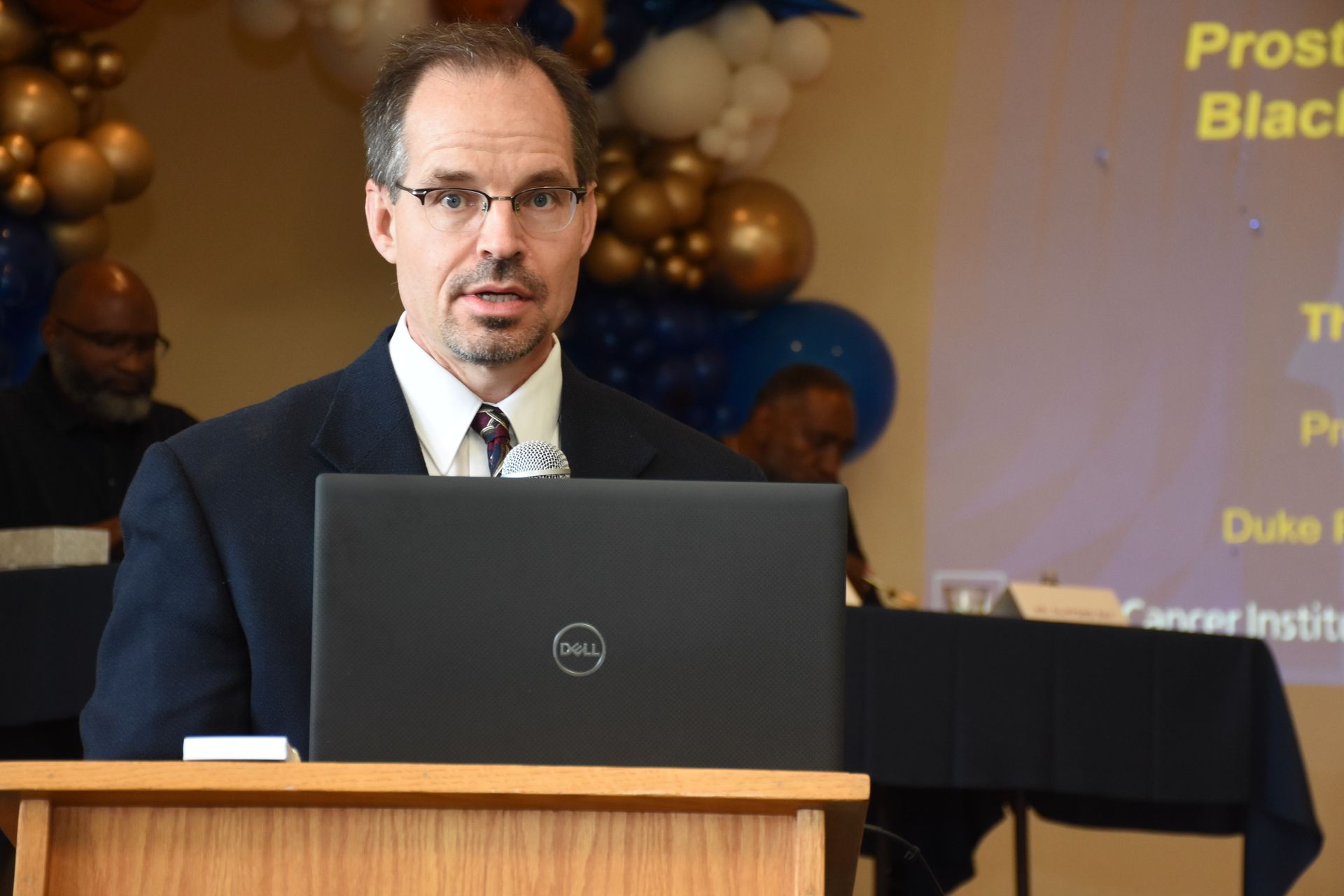 Man speaking at a podium with a laptop, presentation screen in background.
