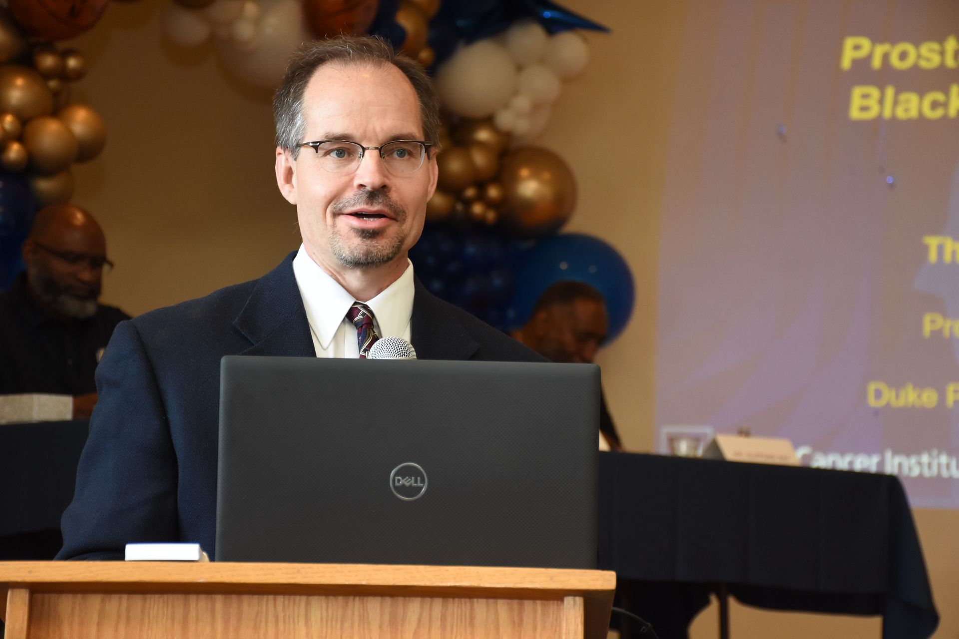 Man speaking at a podium with a laptop, presentation backdrop, and balloons.