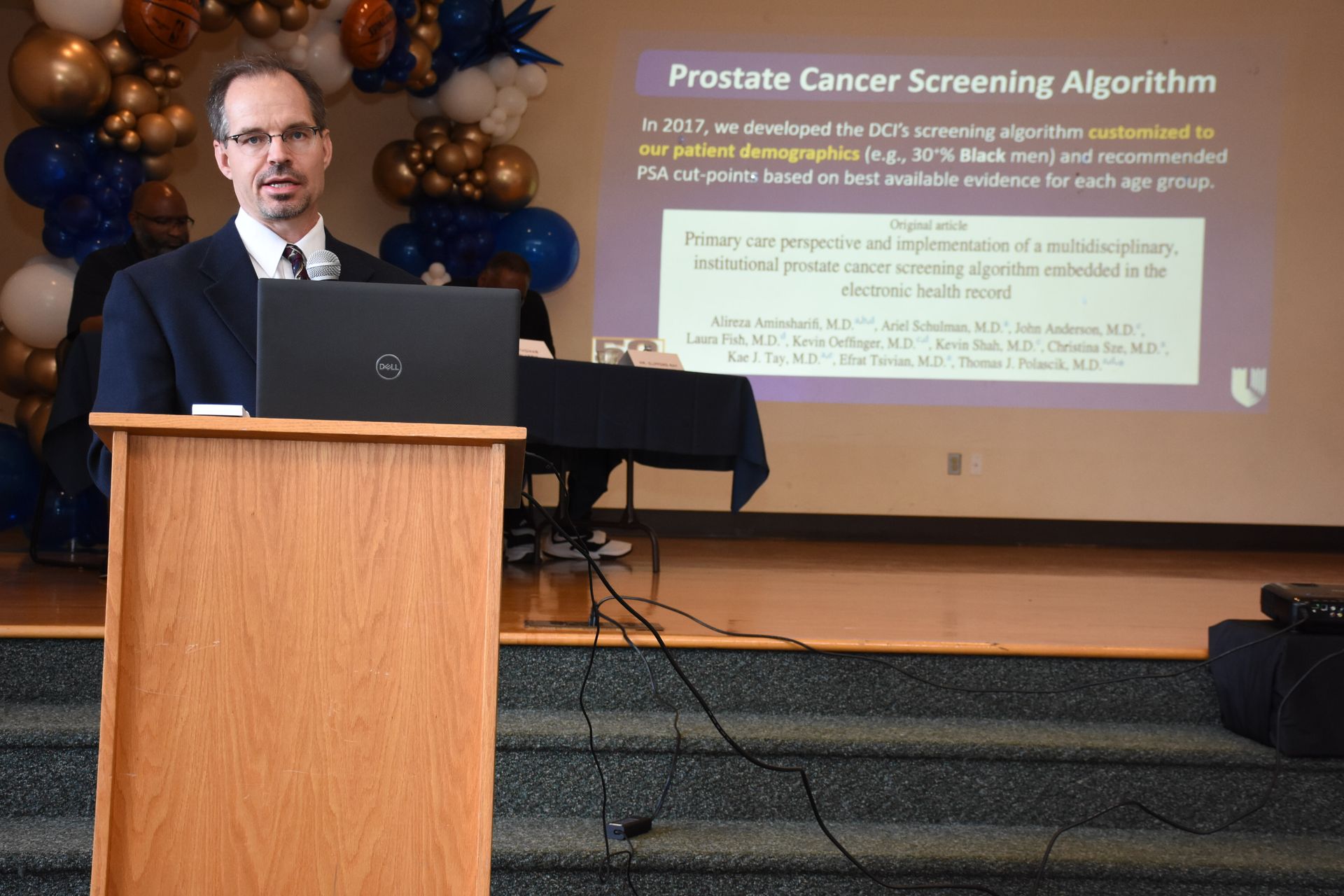 Man at a podium presenting about prostate cancer screening with a laptop and screen showing an algorithm.