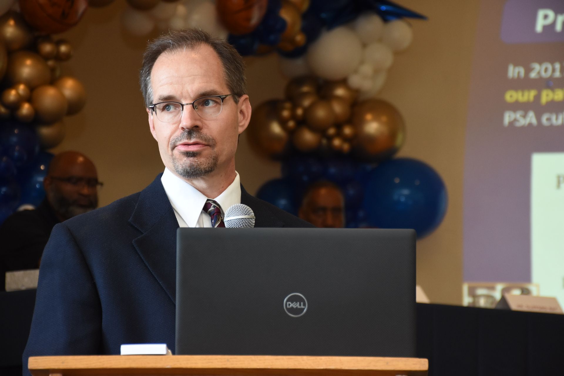 Man in a suit speaks at a podium with a laptop, gold and blue balloon background.