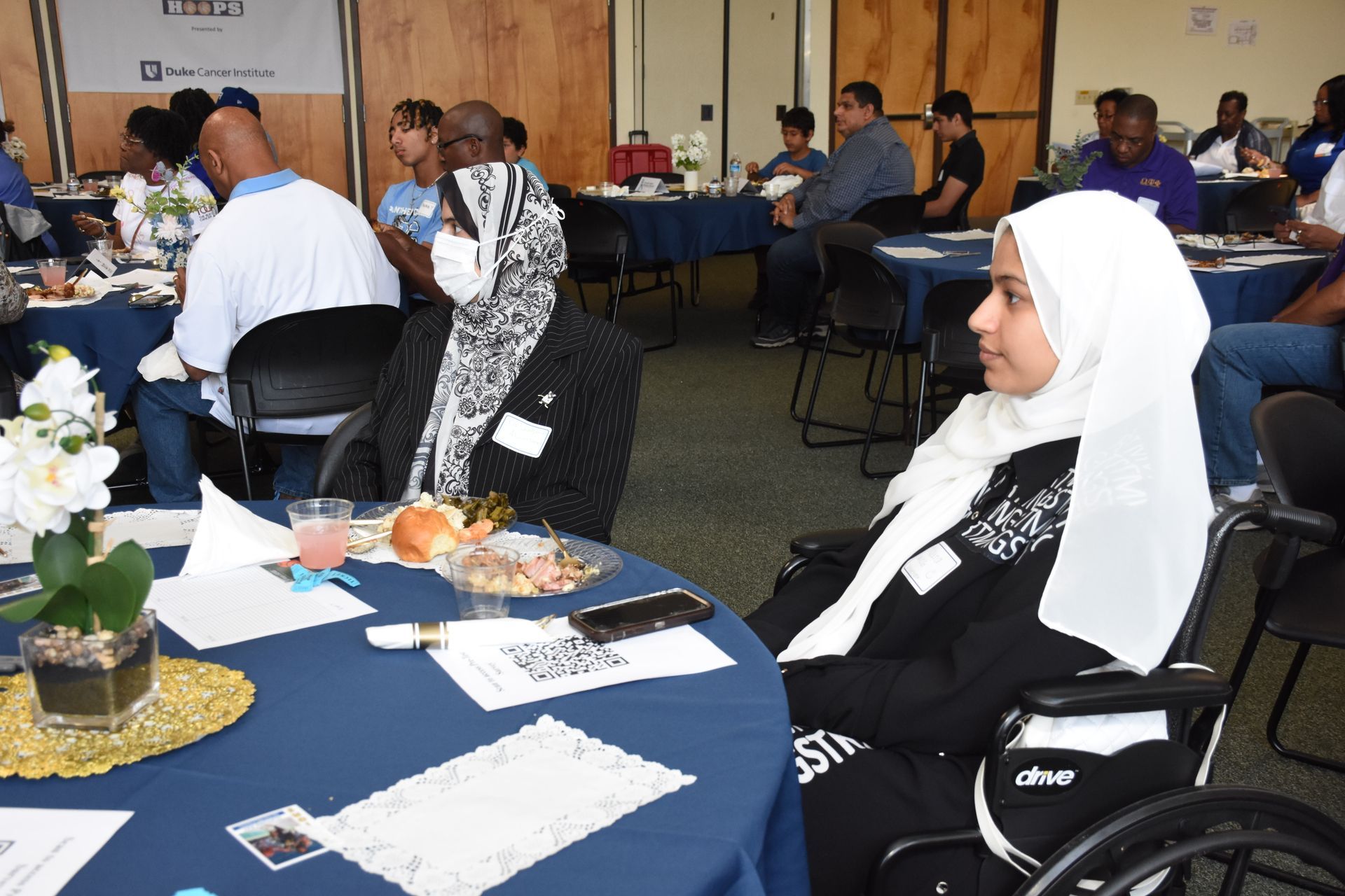 People at a table, one in a wheelchair. Event with blue tablecloths, refreshments, and decor.