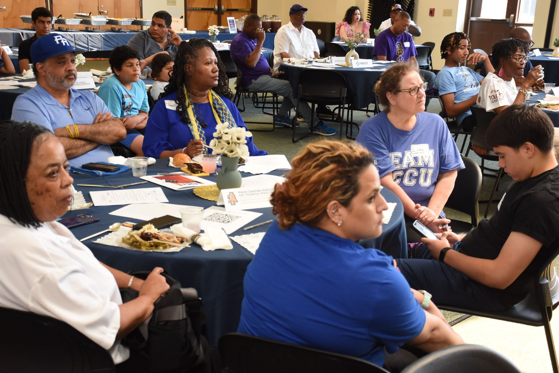 People seated at tables listening, in a large room. Some wear blue shirts. Tables set with food and centerpieces.
