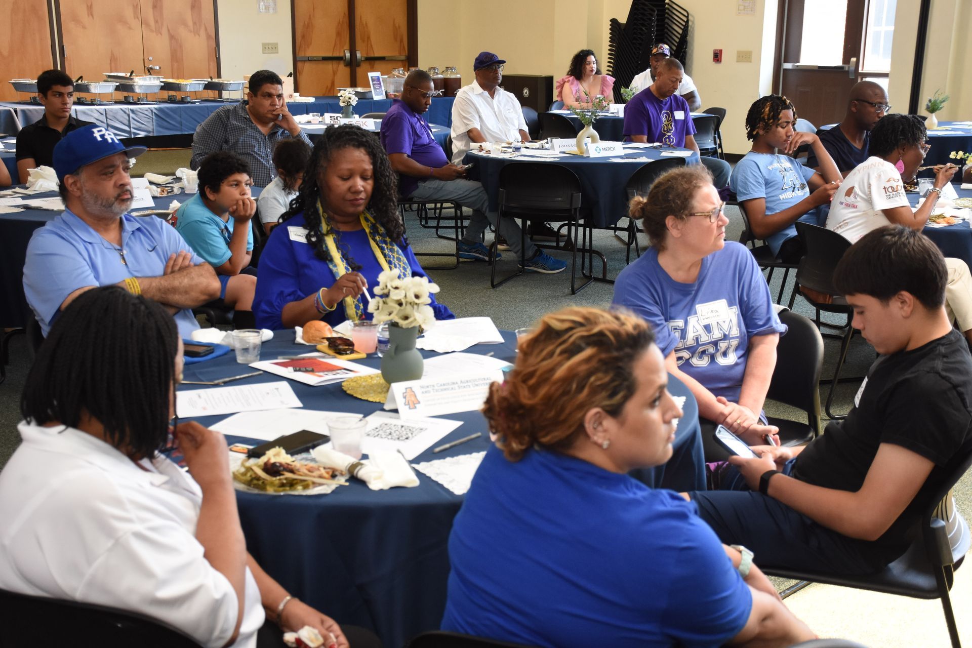 People seated at tables in a room, some wearing blue shirts, engaged in a gathering, possibly a meeting.