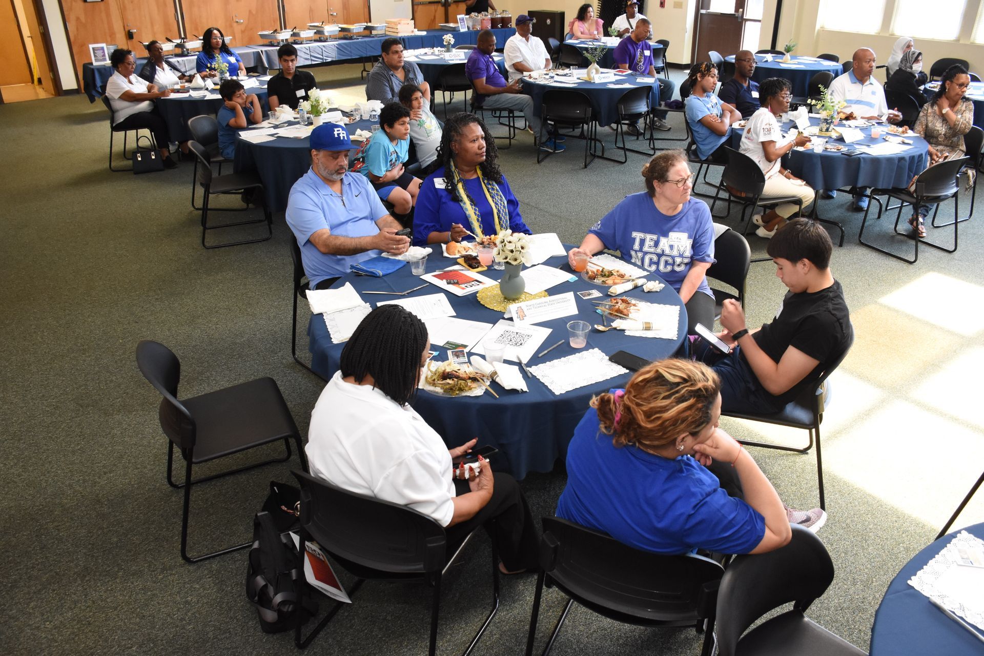 People seated at round tables in a room, some conversing. Tables have blue tablecloths and food.