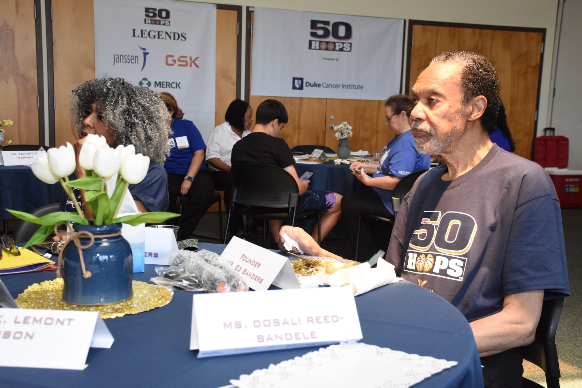 Man at an event with others. Table with flowers, signs, and event branding in the background.