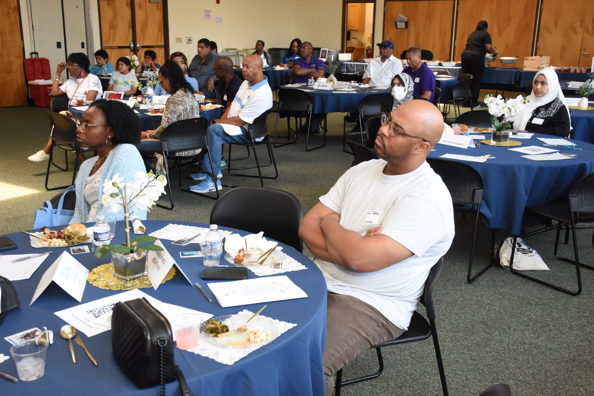 Attendees seated at tables in a room with blue tablecloths, listening attentively. Some have food and water.