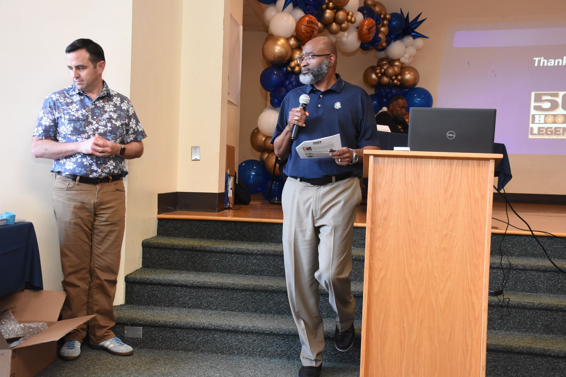 Man speaking at podium, another man stands beside him. Event setting with balloons and laptop.