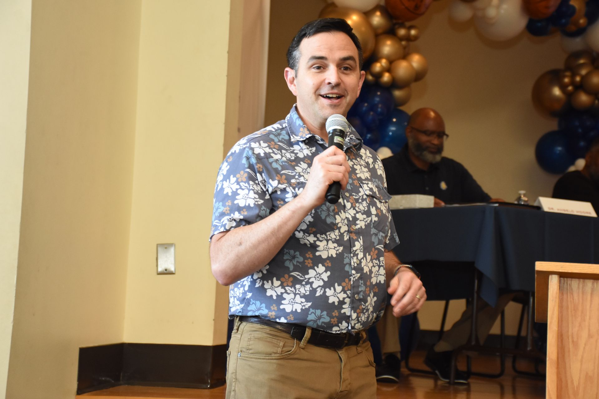 Man speaking into a microphone. He wears a floral shirt and khaki pants at an event with balloons and a table behind him.