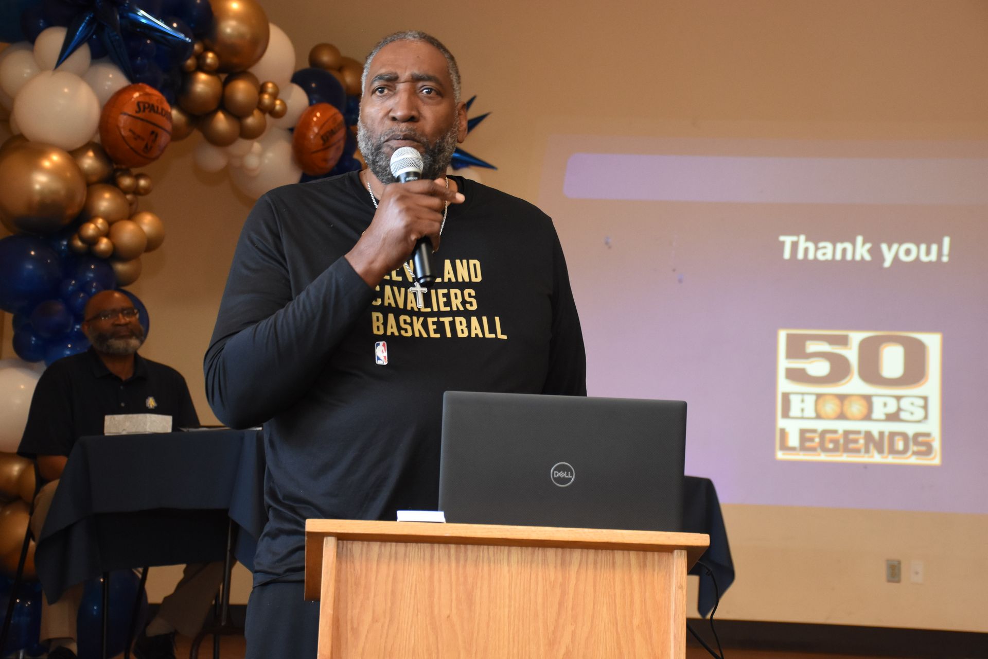 Man speaking into a microphone at a podium with a laptop; balloons and a 