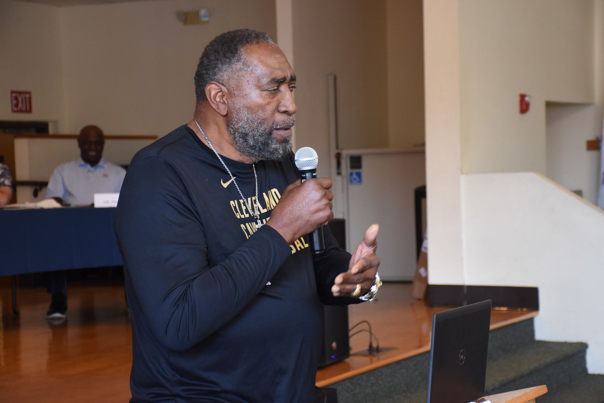 Man speaking into a microphone. He is wearing a black shirt and standing in a room with a laptop.