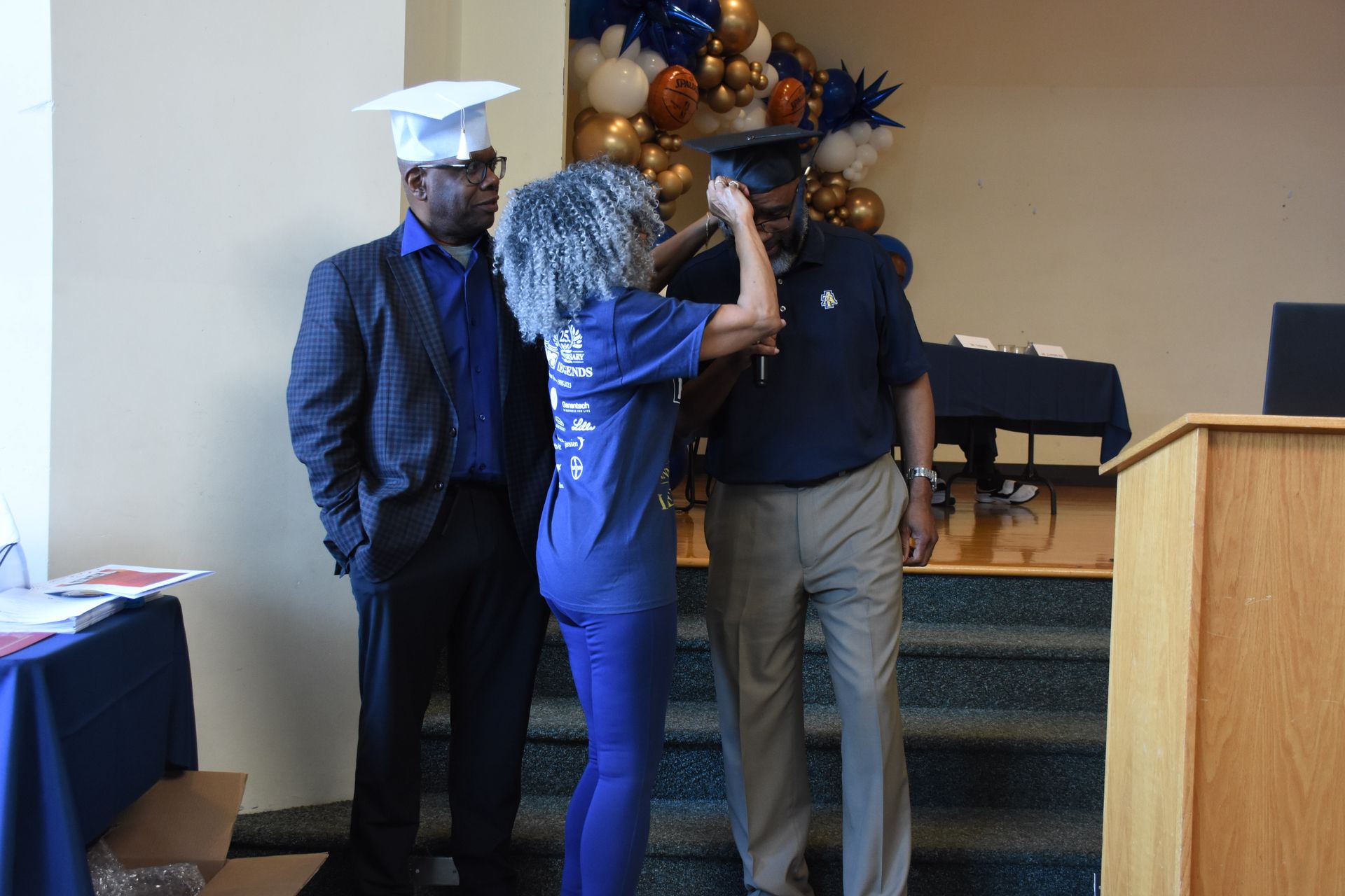 Woman adjusts cap on man's head at graduation. Another man stands nearby. Festive balloons in the background.