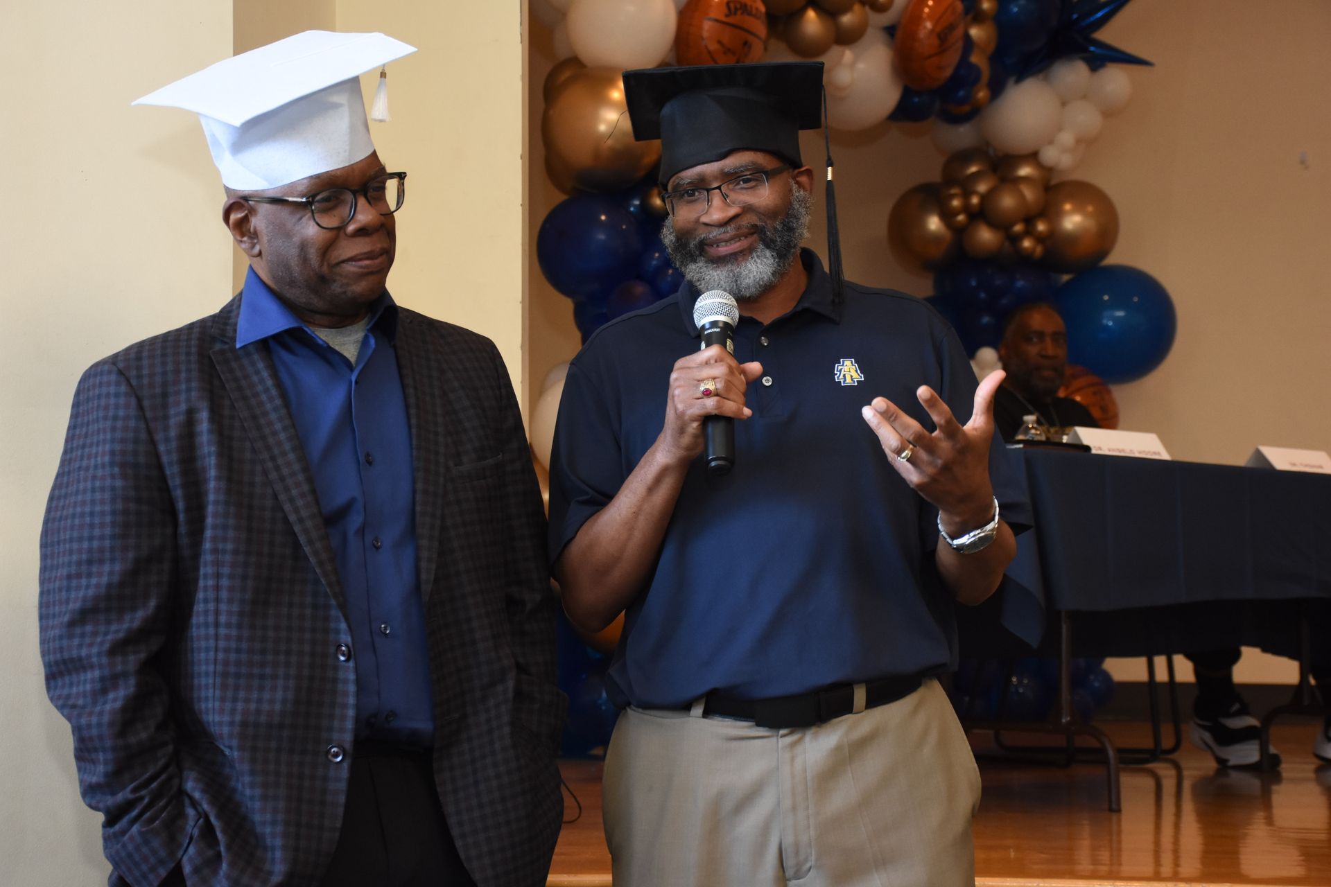 Two men wearing graduation caps, one speaking into a microphone. Balloons in the background.