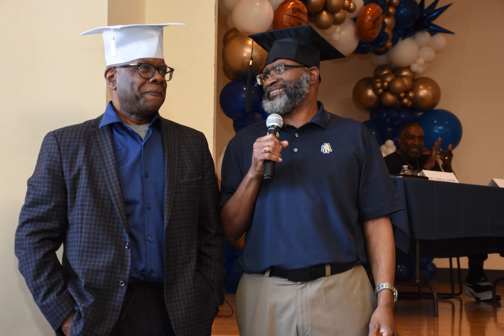 Two men in graduation caps at podium, one speaking into mic. Balloons in background.