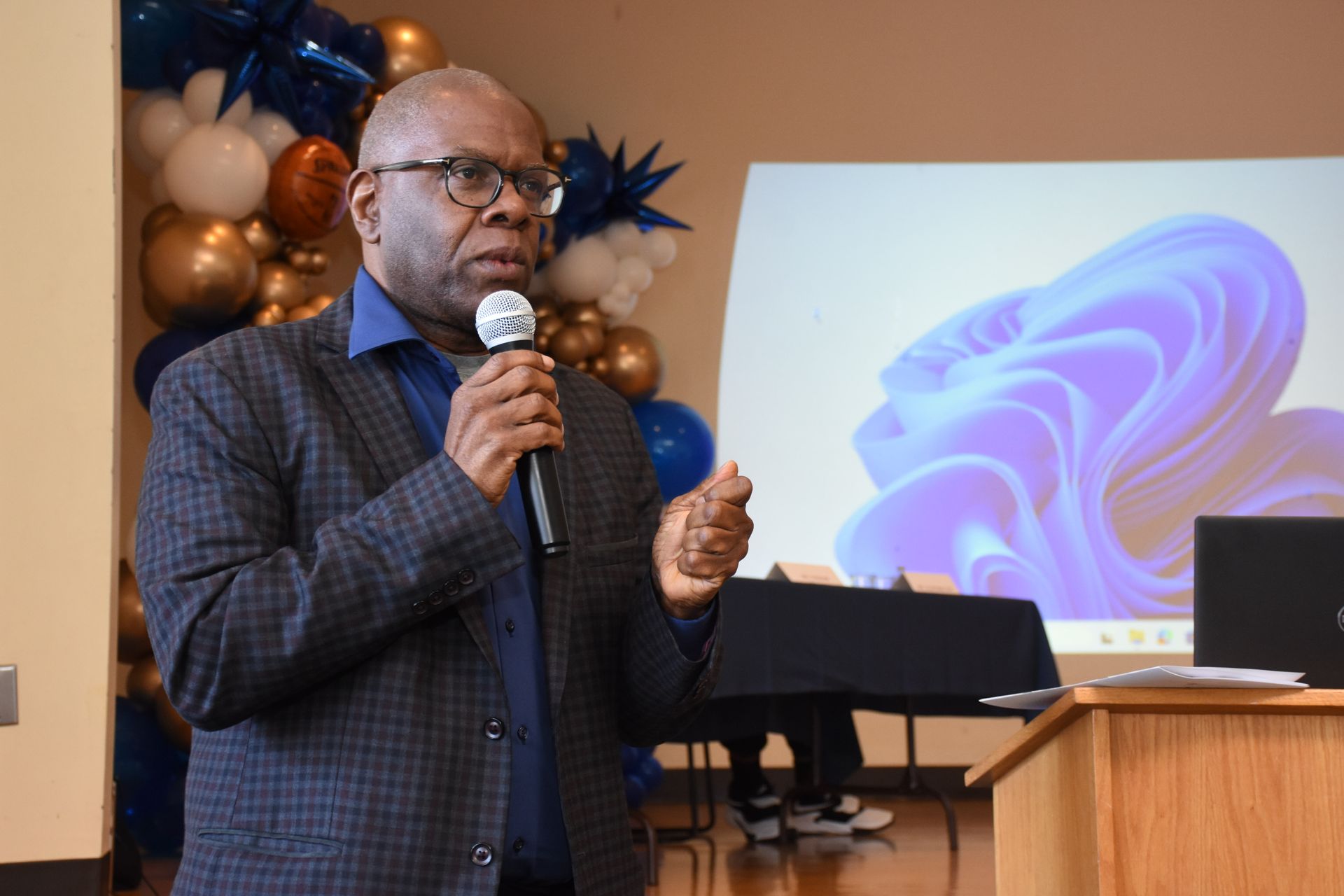 Man speaking into a microphone at an event, with a screen and balloons in the background.